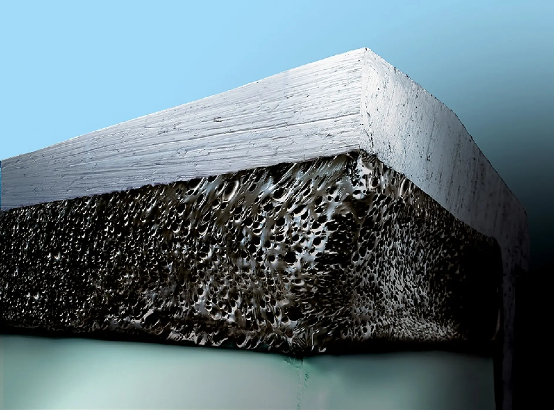 Close-up of a cross-section of a metal block against a blue background. The top layer is smooth, silver, and reflective, while the bottom layer is rough, porous, and dark with a textured surface.