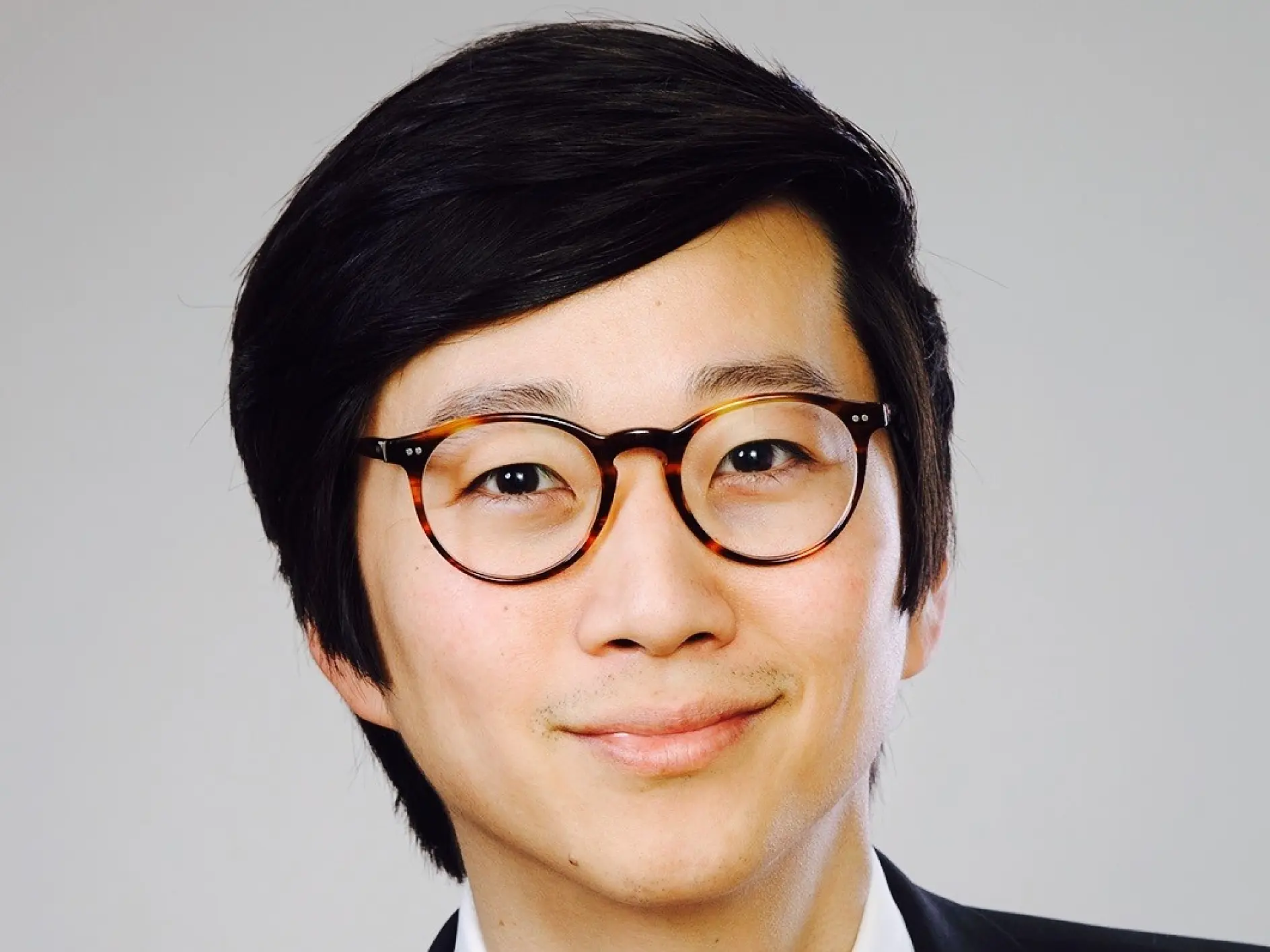 A person with short dark hair wearing round eyeglasses and a suit jacket is smiling against a plain background, using tesa tape.