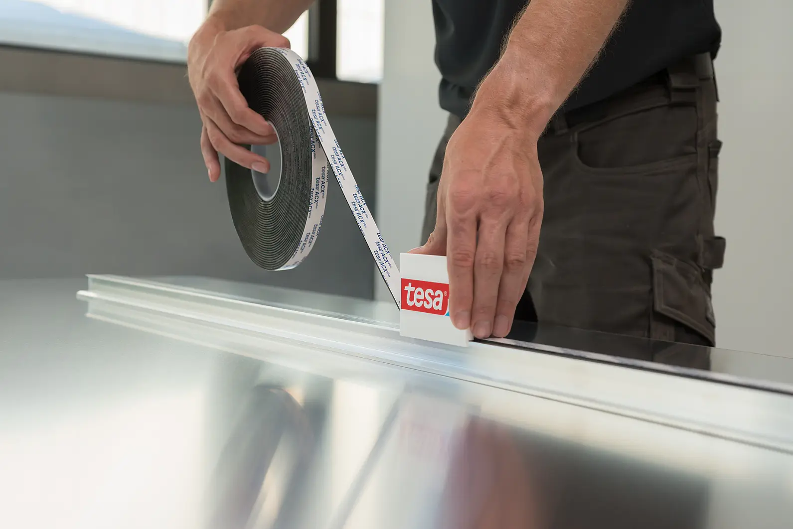 A person applies tesa tape from a roll onto a long, flat surface. The tesa tape roll has a label with the word tesa. The person is wearing a dark shirt and brown pants, and the surface appears to be metallic. (This text has been generated by AI)