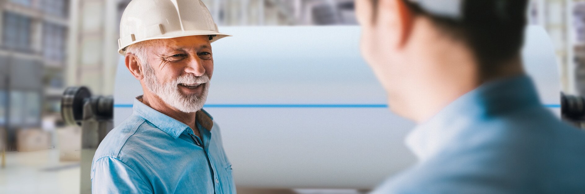 Two men wearing safety helmets shake hands in an industrial setting. One holds a tablet displaying graphs. Large machinery is blurred in the background, suggesting a factory or manufacturing environment, where tesa tape might be commonly used for various applications. (This text has been generated by AI)