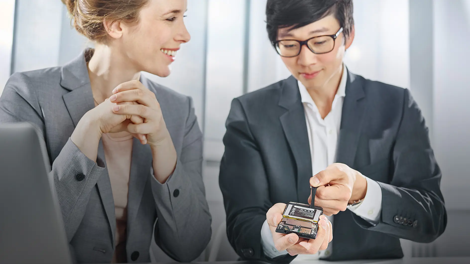 Two people in business attire are seated indoors. One person is holding a small electronic device and tool, gesturing towards it with tesa tape, while the other smiles and observes. A laptop is partially visible on the table. Large windows are in the background.