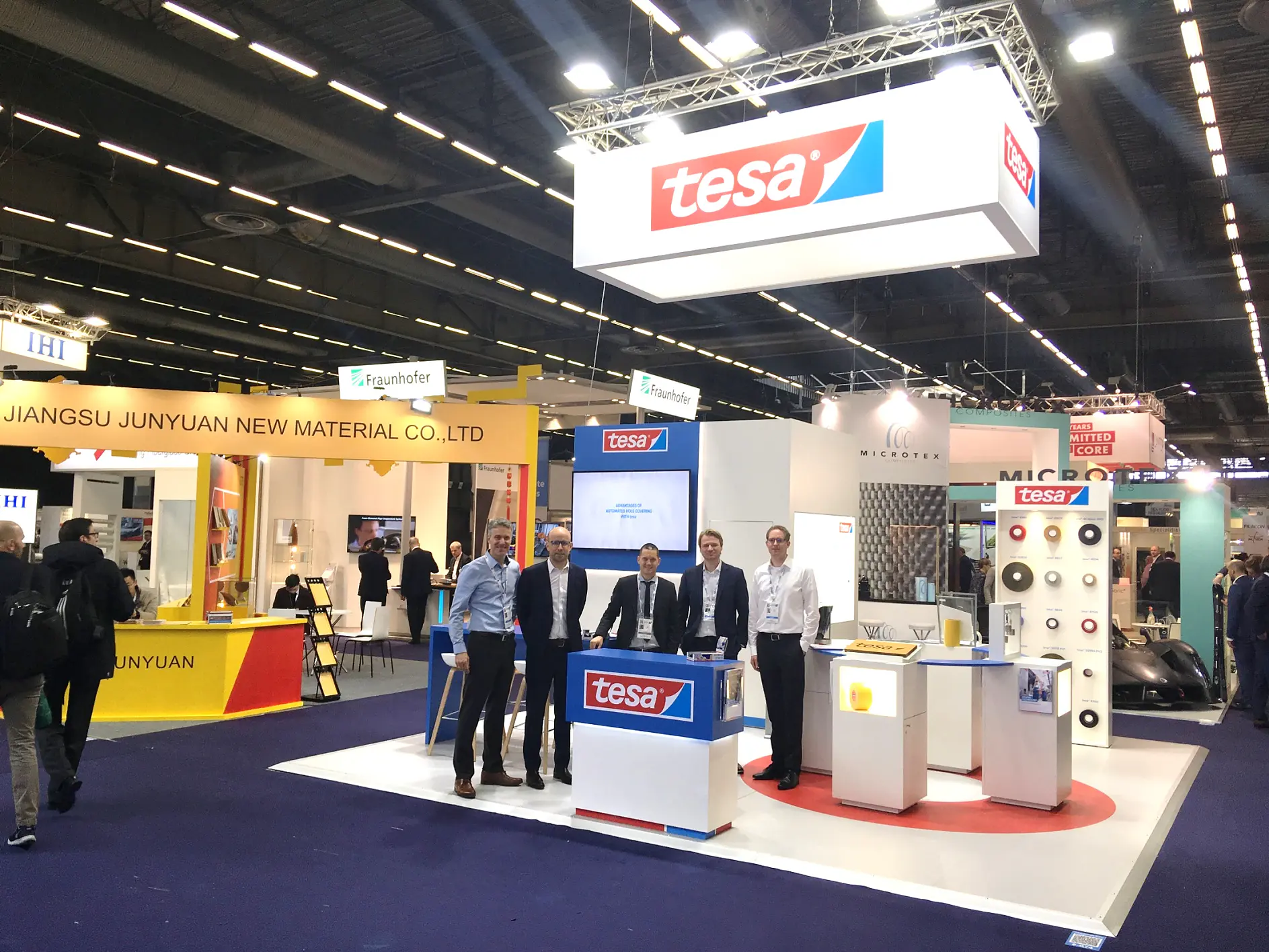 A trade show booth displays the logo tesa prominently. Five people stand in front of the booth, smiling at the camera. Other booths and banners are visible in the background, showcasing various company names and logos.