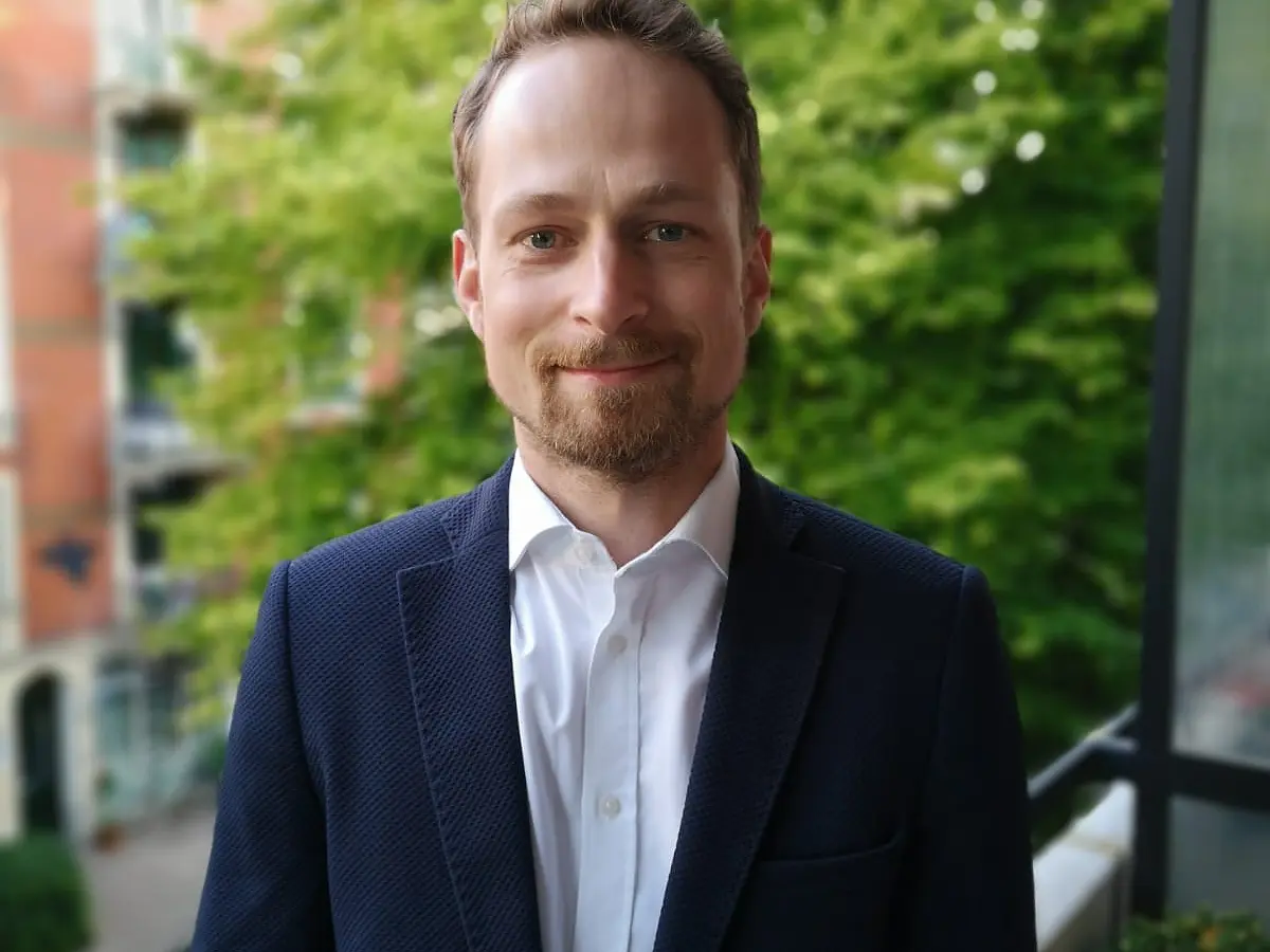 A man with a beard is smiling while standing outside. He is wearing a dark suit jacket over a white shirt. In the background, there are trees and a red brick building.