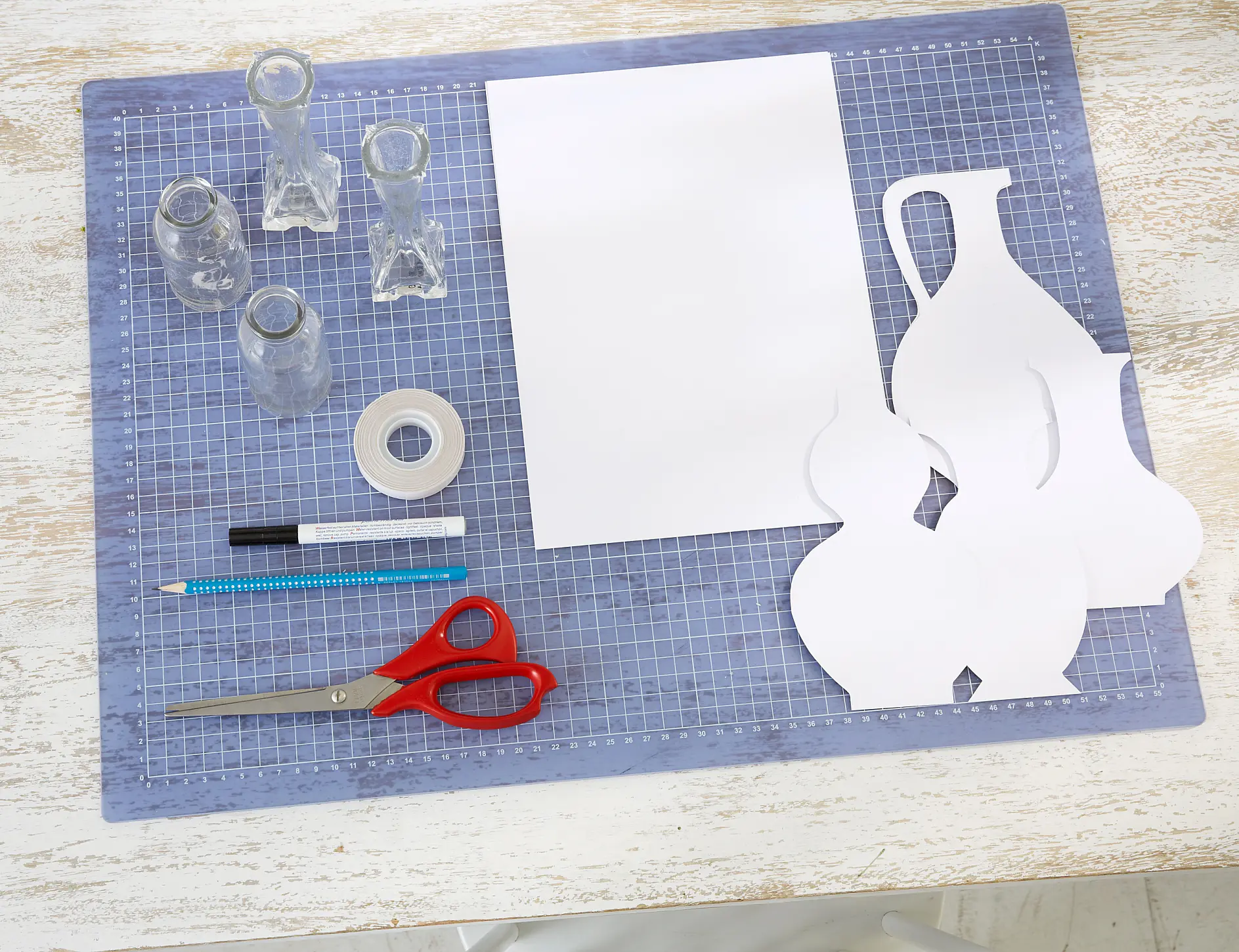 A crafting setup on a table includes a cutting mat with a sheet of white paper, paper vase cutouts, clear tesa tape, red scissors, a black pen, and a blue pencil. Three small glass vases are on the left side.
