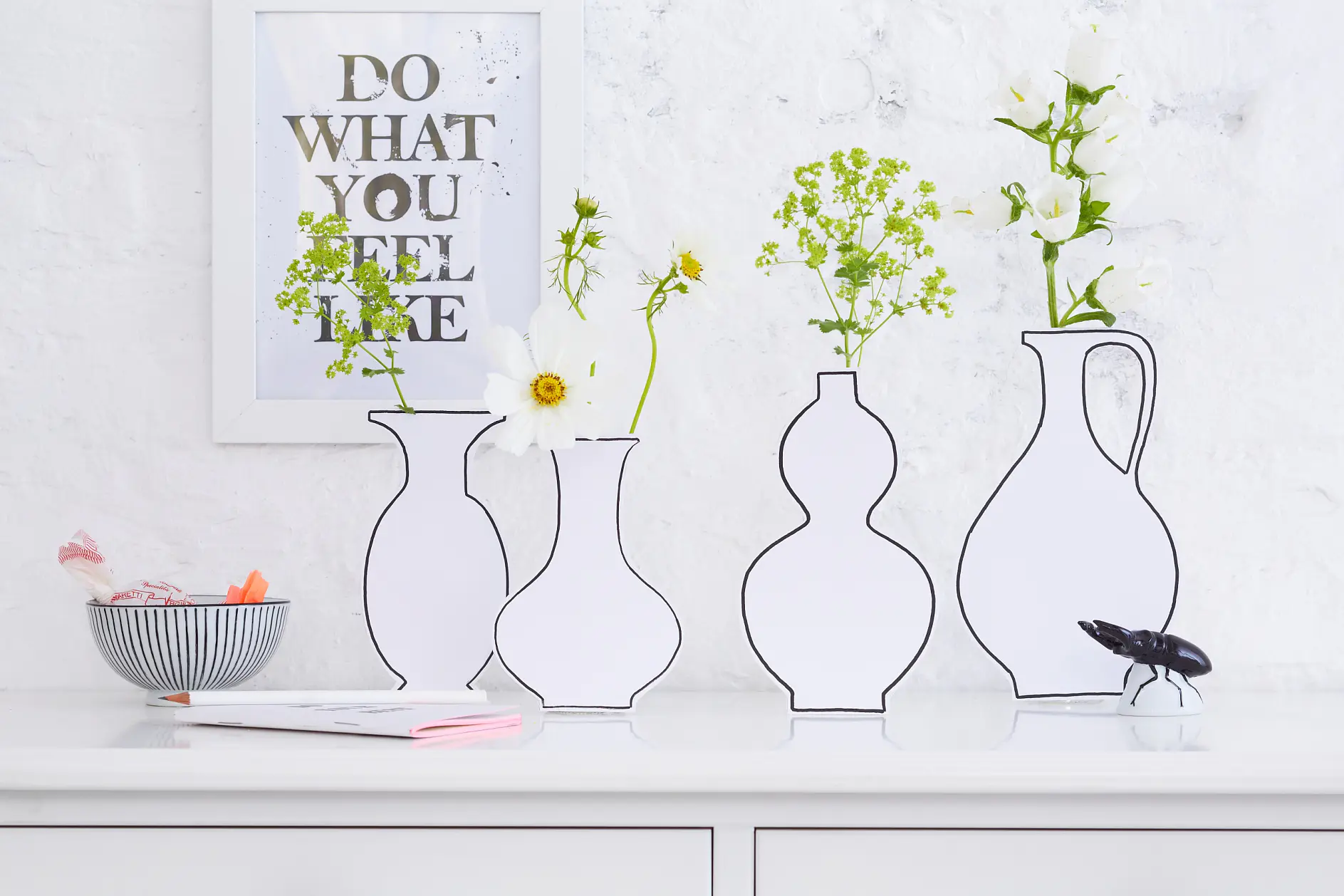 A minimalist arrangement features white vases outlined with black, each holding white flowers and greenery. A framed text saying DO WHAT YOU FEEL LIKE is on a white brick wall. A bowl and pen are on a white surface nearby, secured neatly using tesa tape to maintain the sleek aesthetic of the space.