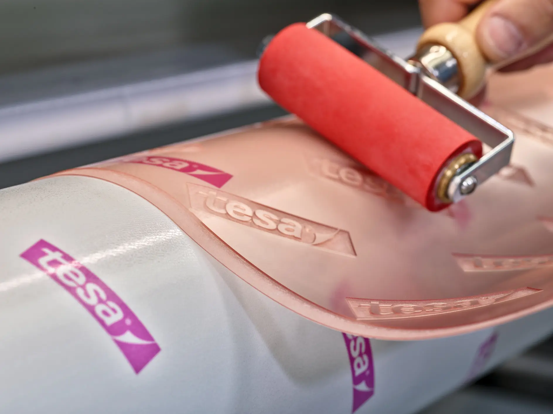 Close-up of a hand using a red roller on a clear adhesive film marked with the tesa tape logo. The film is being applied to a white cylindrical surface.