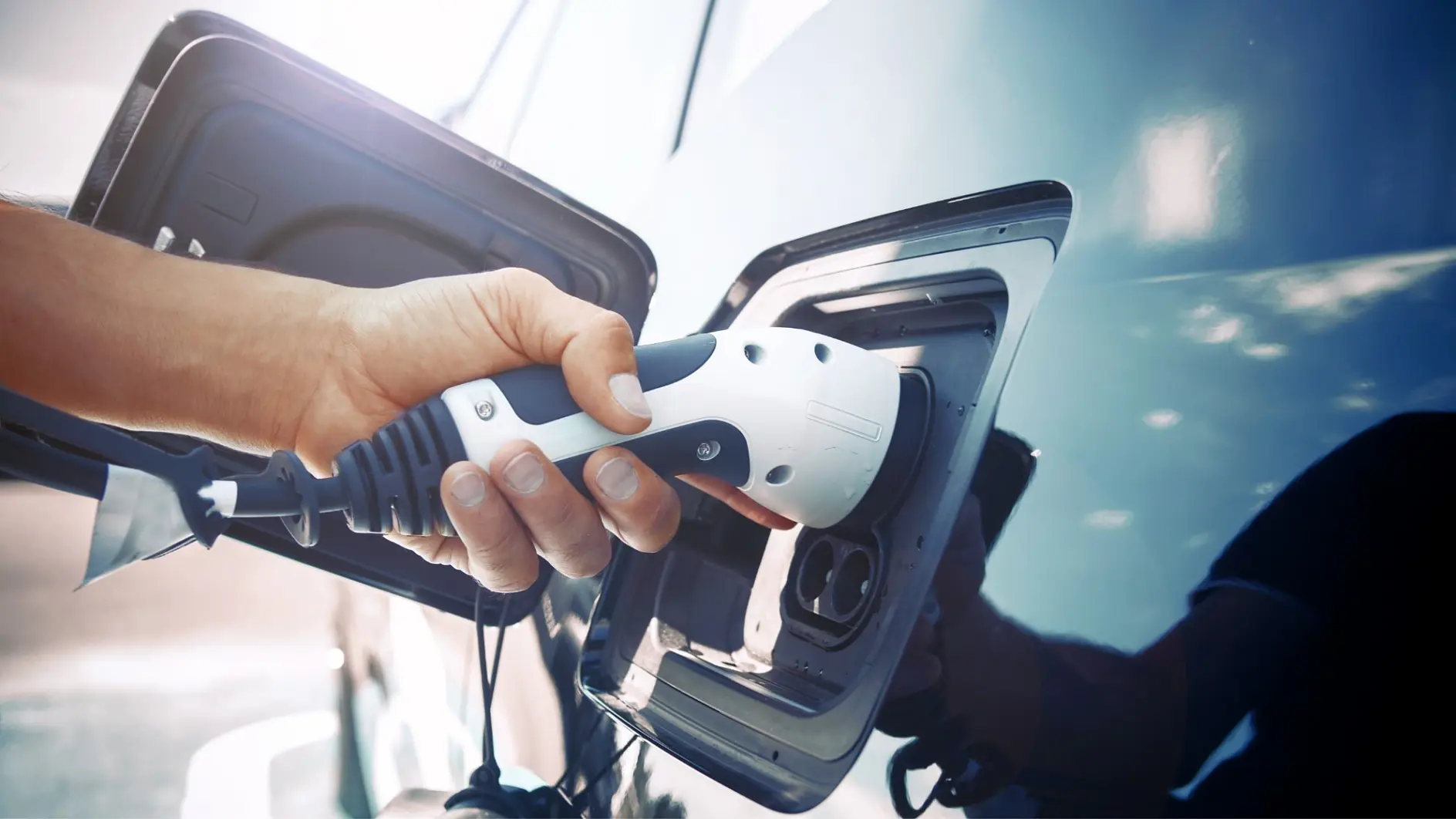 A close-up shows a person plugging a charging cable into an electric vehicle. The hand holds the white connector aligned with the cars charging port, while tesa tape can be seen securing nearby cables. The cars sleek blue exterior is visible in the background.