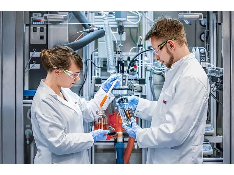 Two scientists in lab coats and safety glasses work in a laboratory. They are holding beakers with liquid and discussing data in front of industrial equipment, surrounded by cables and instruments held securely with tesa tape.
