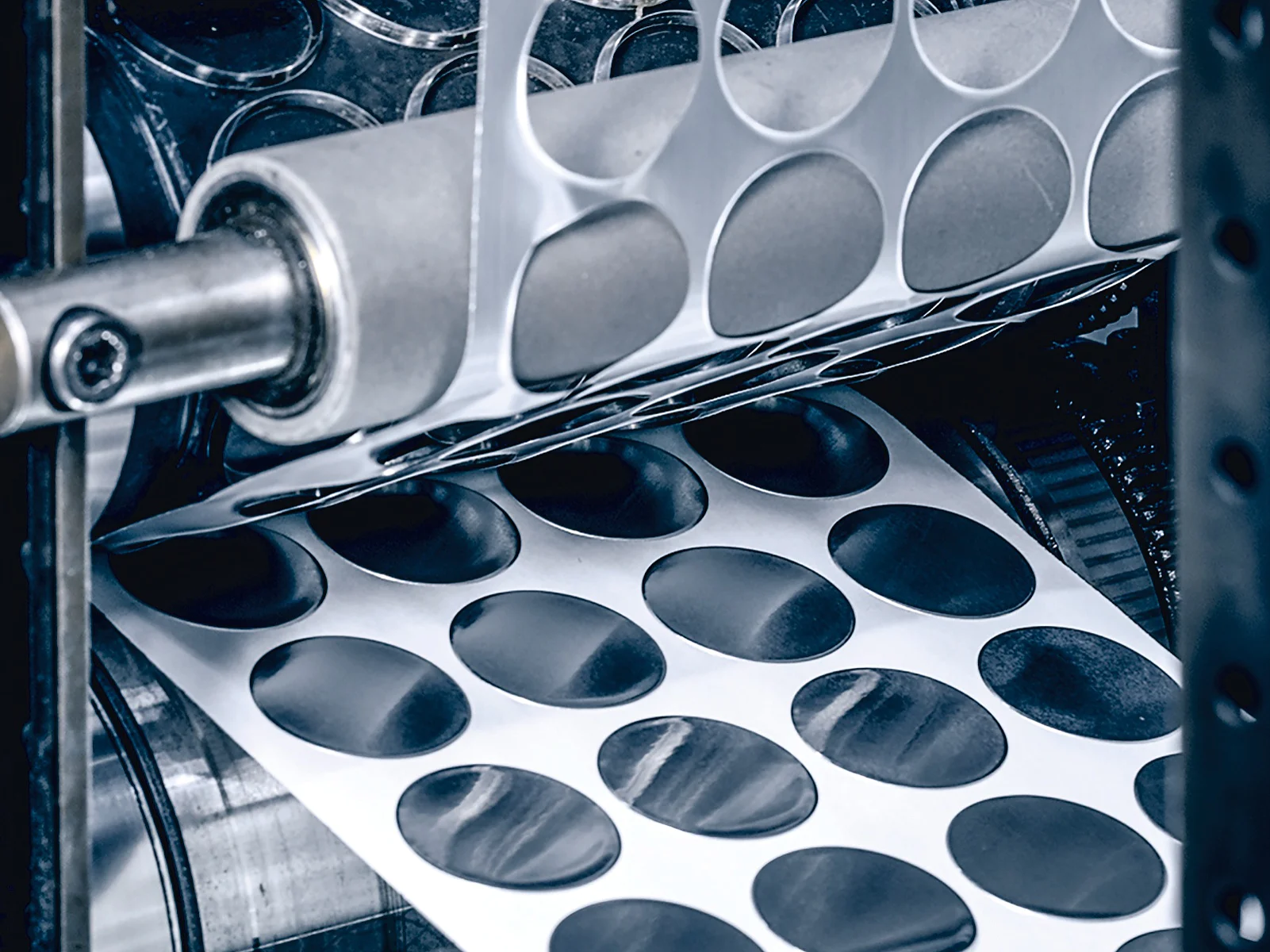 A machine with rollers processes sheets of adhesive paper, cutting out circular stickers. Several rows of stickers are visible, partially separated from the sheet. The surrounding machinery is metallic, with visible gears and supports showcasing tesa tape in action. (This text has been generated by AI)