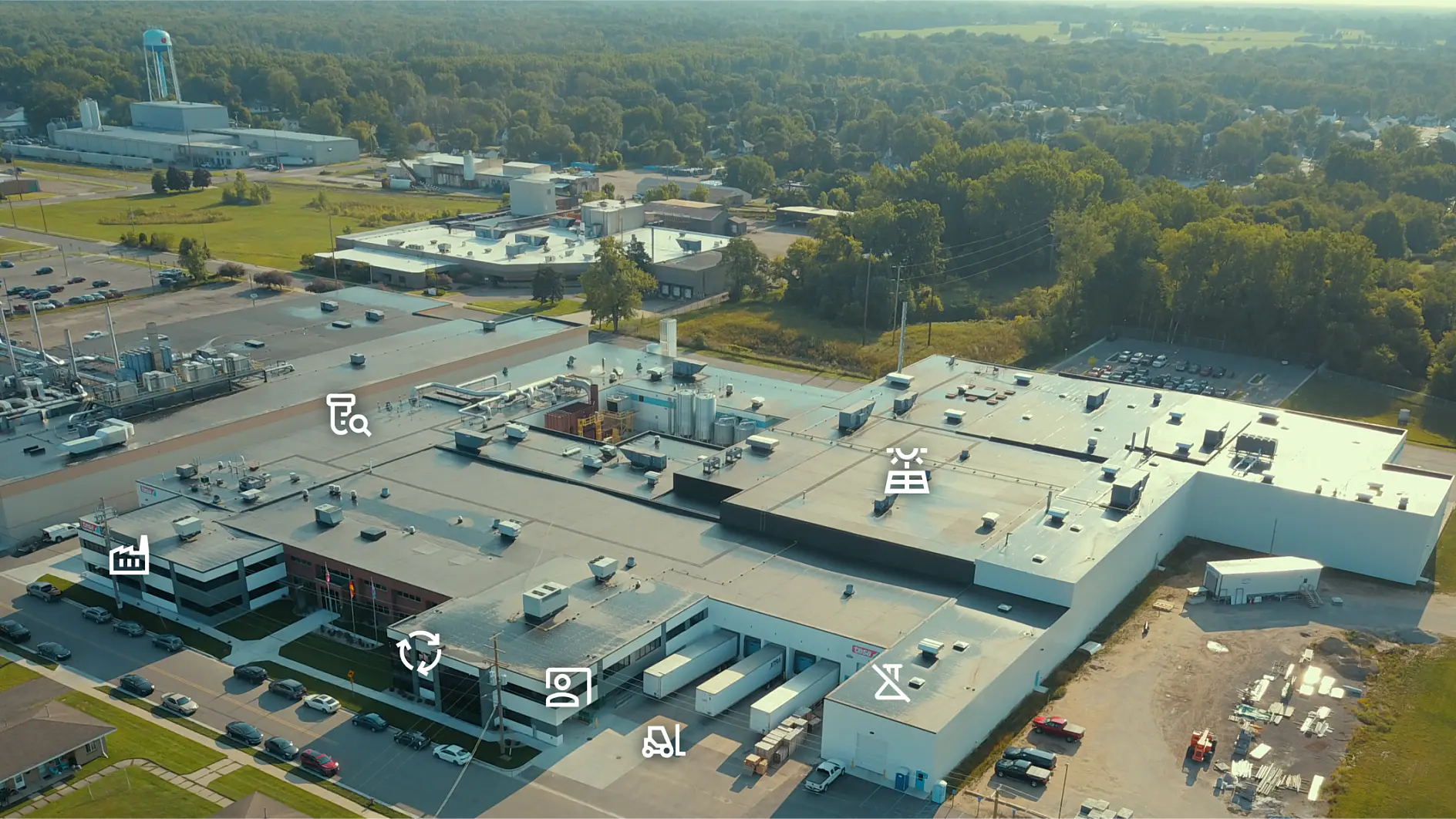 Aerial view of an industrial complex with multiple large buildings surrounded by parking lots and green areas. Various industrial icons are overlaid on the image, indicating different sectors or components within the facility. (This text has been generated by AI)