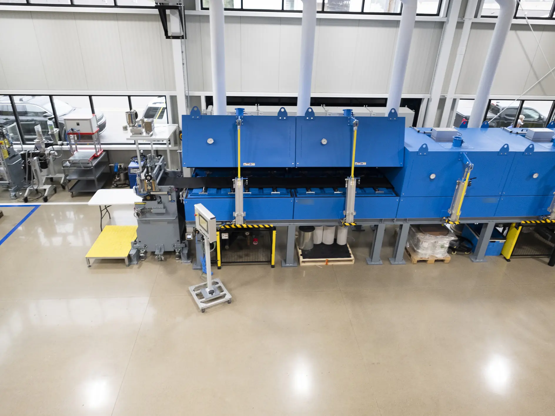 A large industrial machine with a blue exterior is positioned on a clean factory floor. Several pipes and components are attached using tesa tape. The area is well-lit with windows above. Nearby are tables, boxes, and other equipment secured or labeled with tesa tape. (This text has been generated by AI)