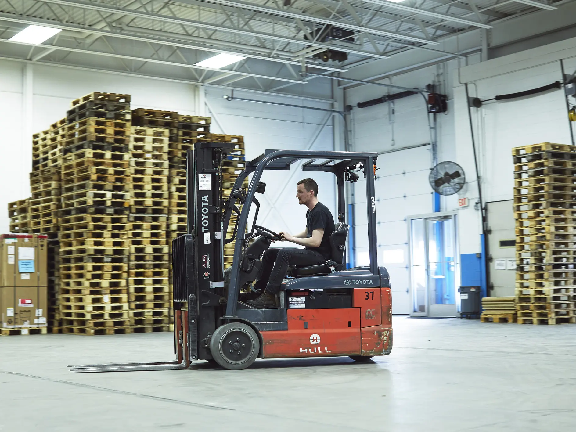 A person operates a red Toyota forklift inside a warehouse. Stacks of wooden pallets are seen in the background. The warehouse has white walls, a blue section, and several overhead lights. (This text has been generated by AI)
