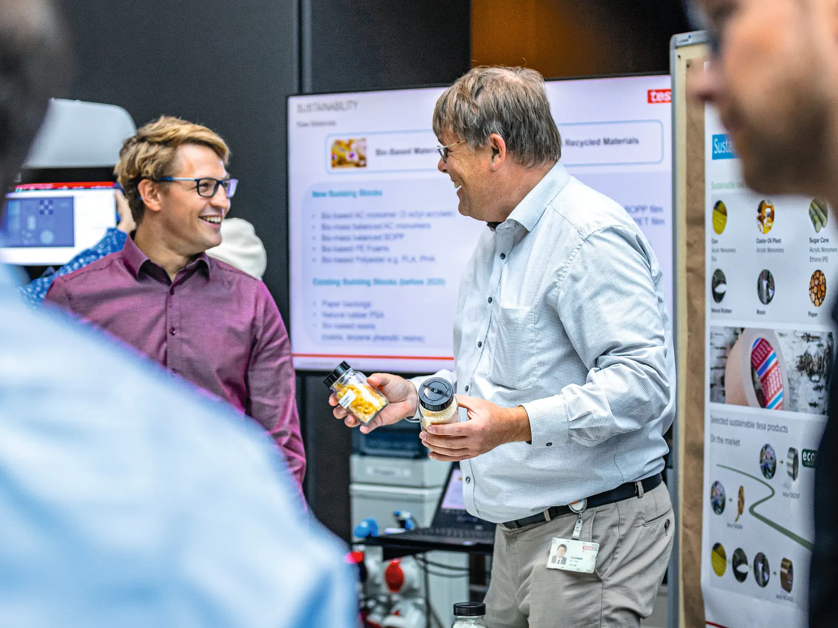 Two men stand at a table with various items and posters, engaging in conversation. One man holds a container made by tesa tape. The background features displays and a screen with text. Others are partially visible in the foreground and background. (This text has been generated by AI)