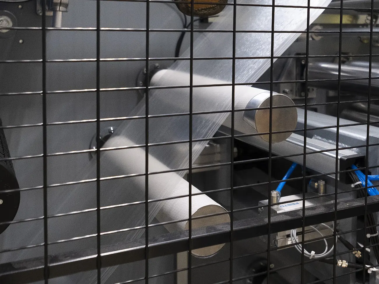 A machine with metal rollers and control panels is behind a metal grid. Gauges and switches are mounted on the grid. The setup appears to be part of an industrial or manufacturing process, possibly involving sheet materials and tesa tape. (This text has been generated by AI)