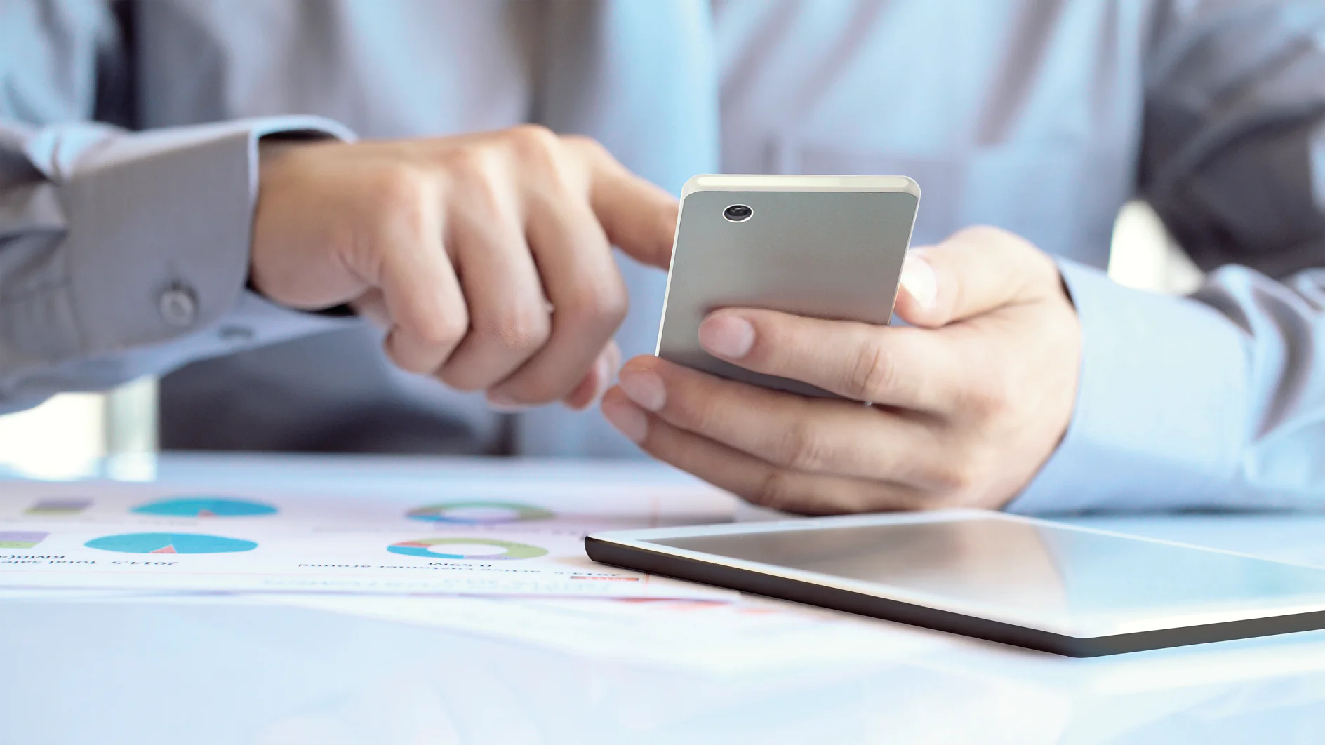 A person in a light blue shirt is using a smartphone, with an open binder or paper featuring colorful charts on the table, secured by tesa tape. Part of a tablet is visible in the foreground, slightly out of focus. The setting suggests a work environment. (This text has been generated by AI)
