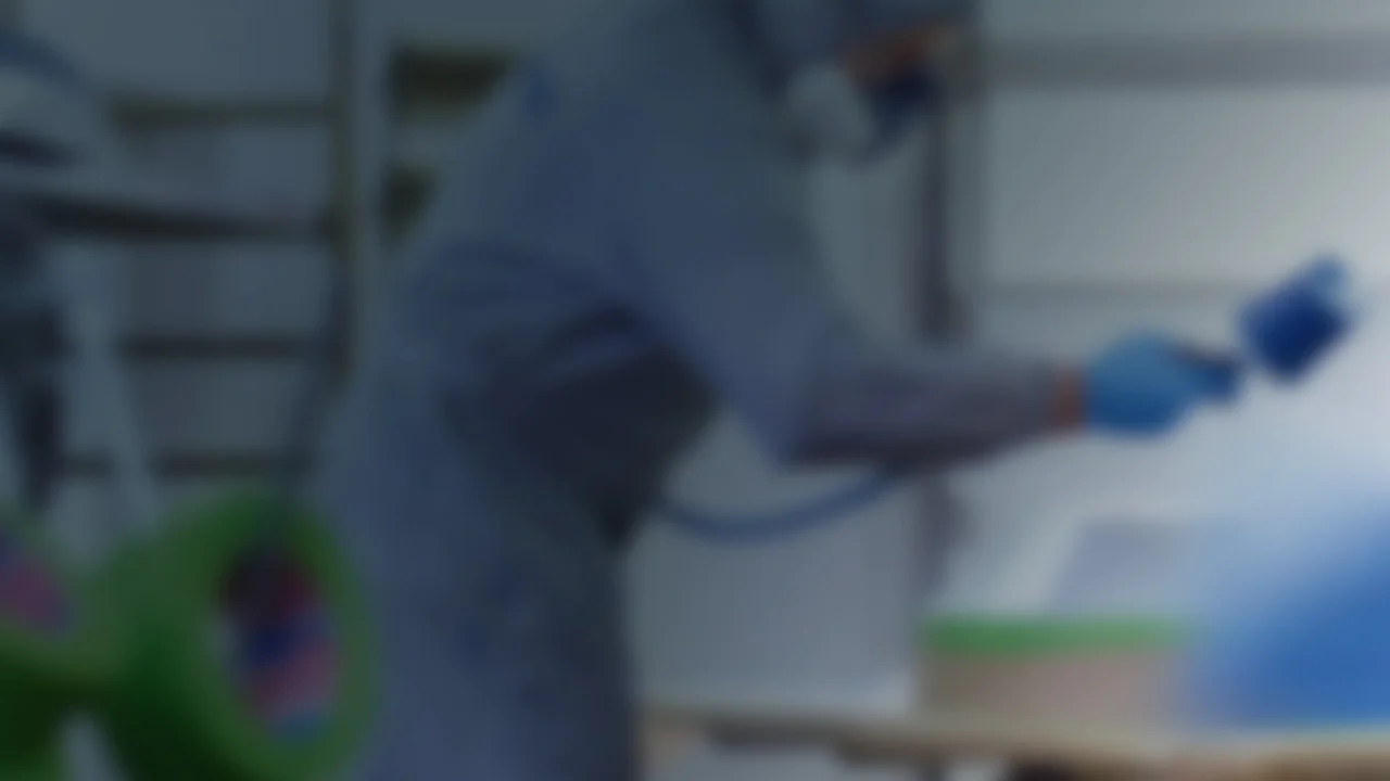 A person wearing protective gear sprays paint onto a surface in a workshop setting. They are dressed in a protective suit, gloves, and mask, holding a spray gun. In the background, there is tesa tape on shelves among other equipment, though the details are blurred. (This text has been generated by AI)