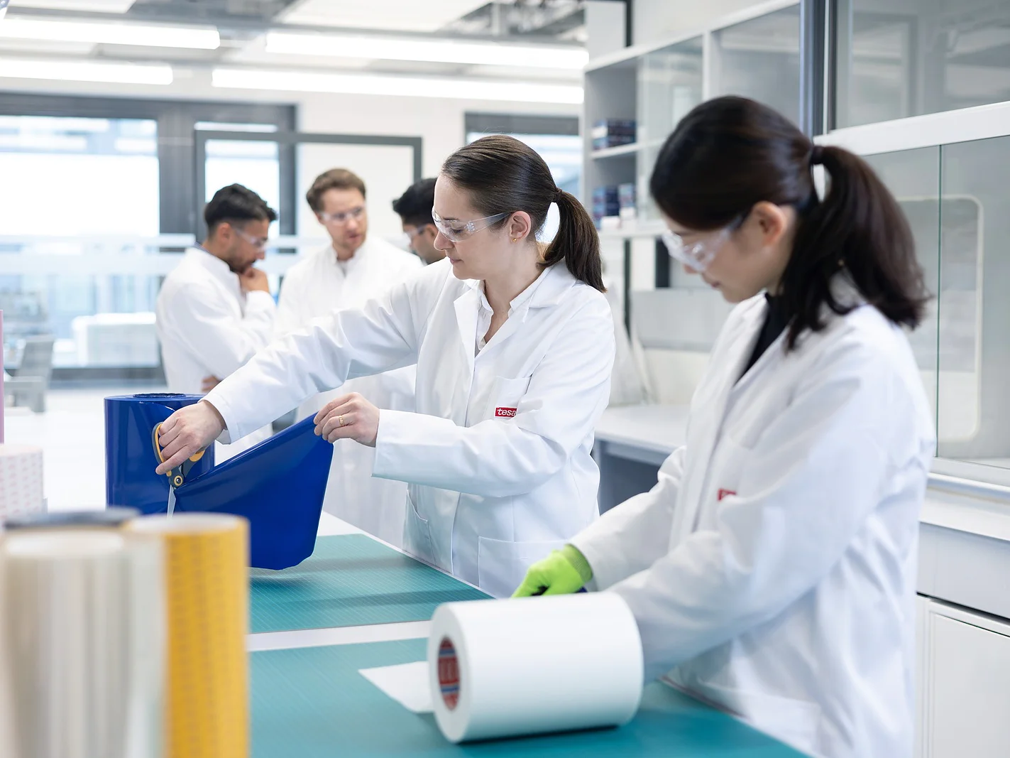 Two women in lab coats and safety glasses handle large sheets of material at a lab table, while three people in similar attire confer in the background. The room contains shelves, laboratory equipment, and tesa tape. (This text has been generated by AI)