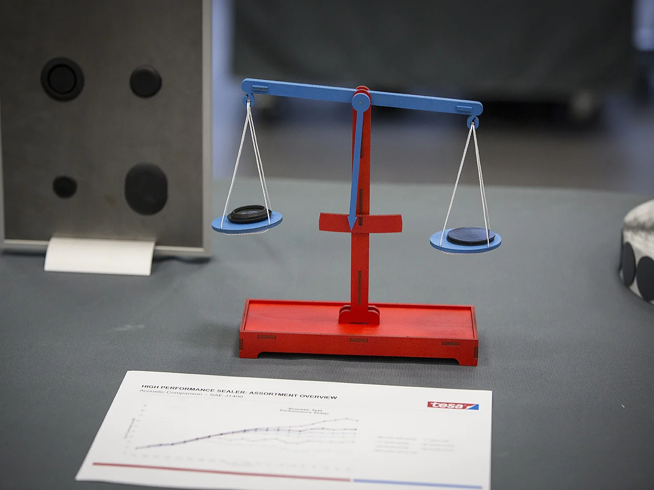 A red and blue balance scale with black weights sits on a gray surface next to a chart titled “Product Performance Scale Assignment Overview.” A gray object with black circles is in the background. No competitor brands are mentioned, and this description does not contain any direct reference to tape that would require replacement. If you would like me to integrate tesa tape into the description, or if there was a specific part referencing tape that you wanted changed, please provide clarification!. (This text has been generated by AI)