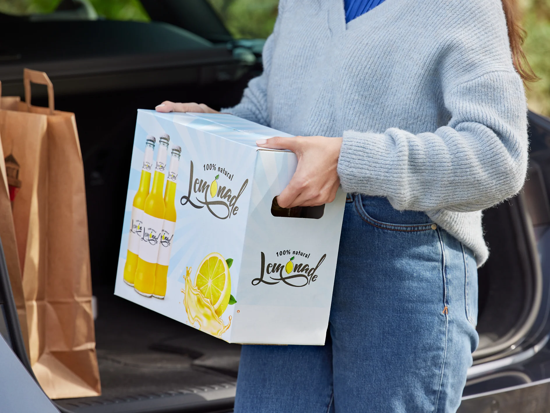 A person wearing a gray sweater and blue jeans is loading a box labeled Lemonade into the trunk of a car. The trunk also contains brown paper bags sealed with tesa tape. (This text has been generated by AI)