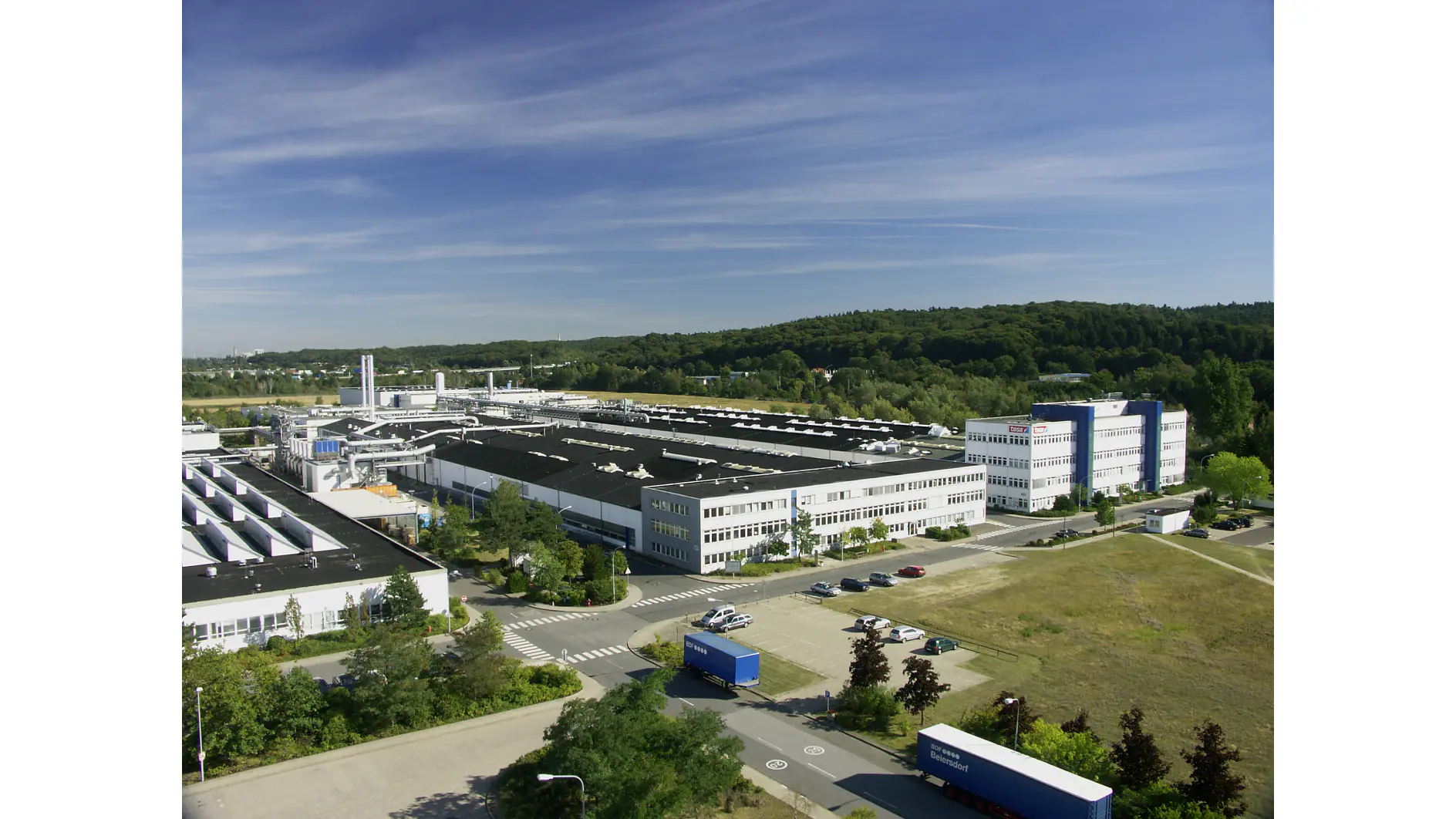 A large industrial complex with multiple white factory buildings and offices is surrounded by greenery, parking areas, and several blue trucks on paved roads under a clear blue sky. The facility specializes in the production and distribution of tesa tape. (This text has been generated by AI)