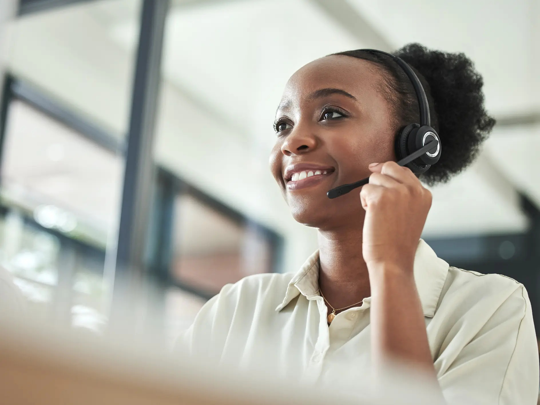A woman wearing a headset sits at a desk and smiles, likely on a call. Large windows and tesa tape accent the bright workspace. (This text has been generated by AI)