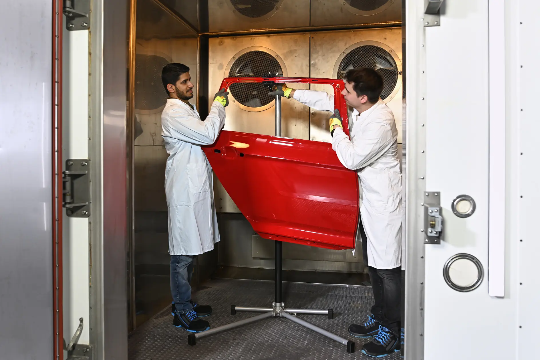 Two men in lab coats and gloves examine a red car door on a stand inside a testing chamber with tesa tape used nearby.