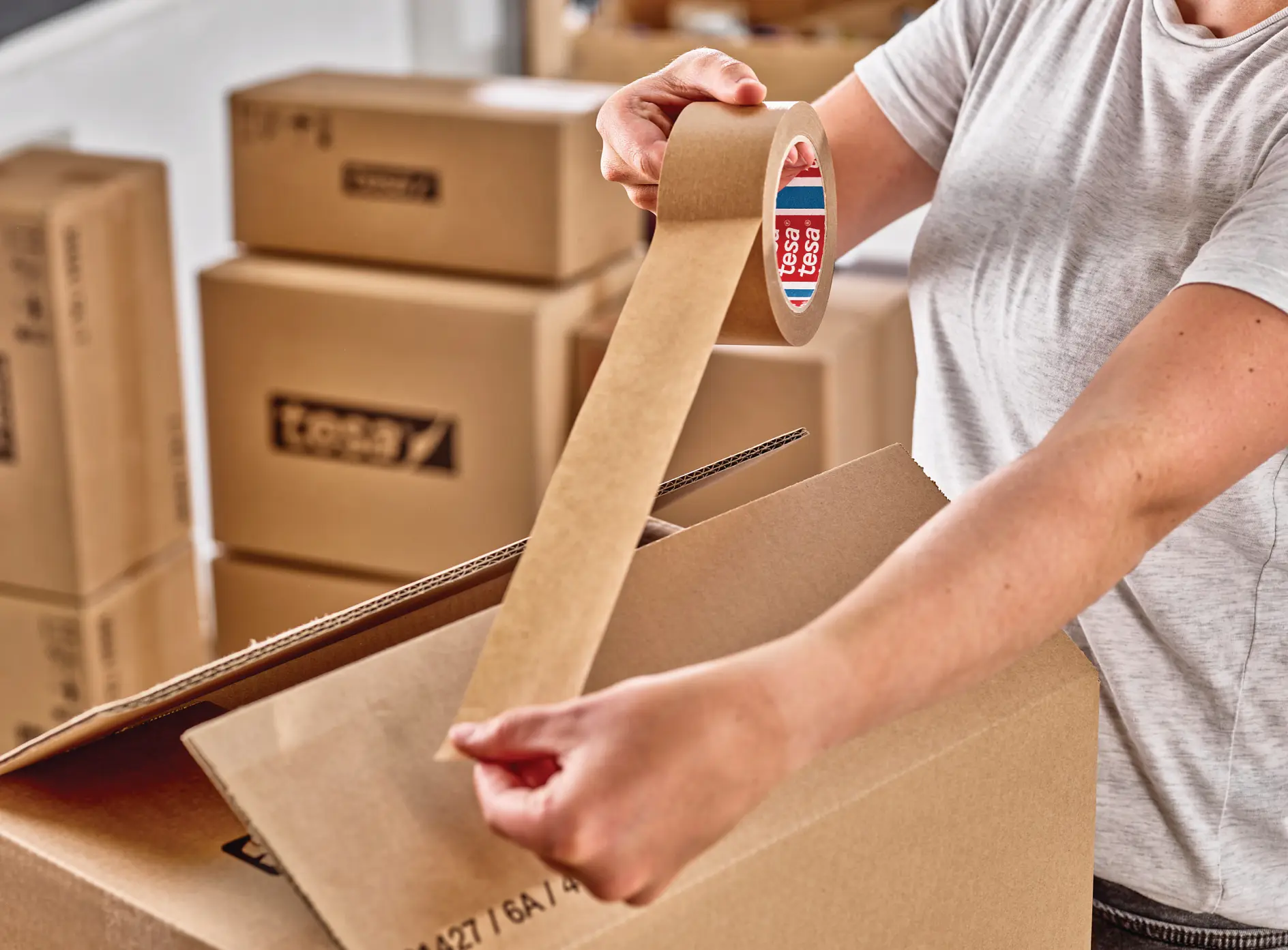 person sealing a cardboard box with brown packing tape in a room filled with stacked moving boxes (This text has been generated by AI)