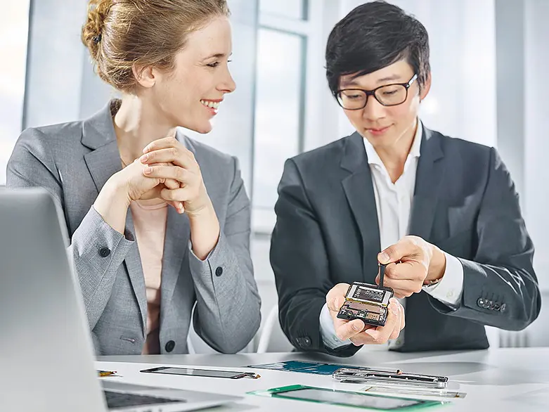 Two business professionals in suits discussing a smartphone at a desk with documents and a laptop in a bright office. (This text has been generated by AI)