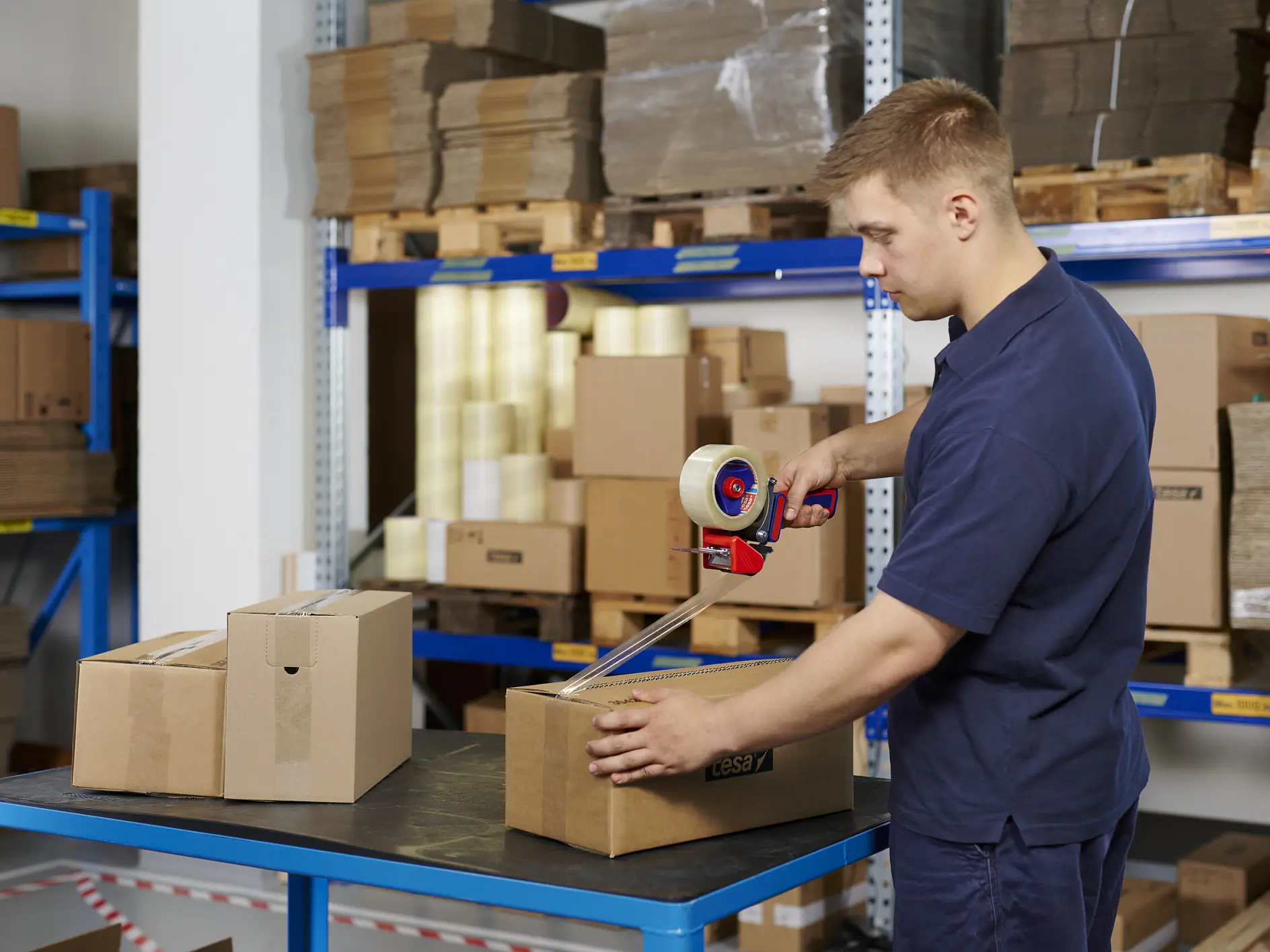 man in navy blue uniform sealing cardboard boxes with packing tape in a warehouse with shelves of stacked boxes and packaging materials (This text has been generated by AI)