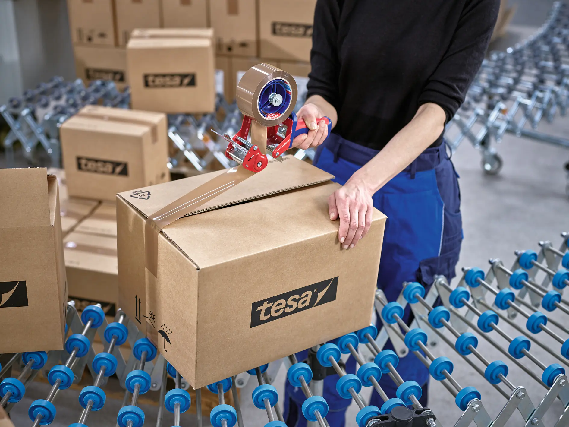 person sealing a tesa-branded cardboard box with tape dispenser on a conveyor belt in a warehouse setting (This text has been generated by AI)