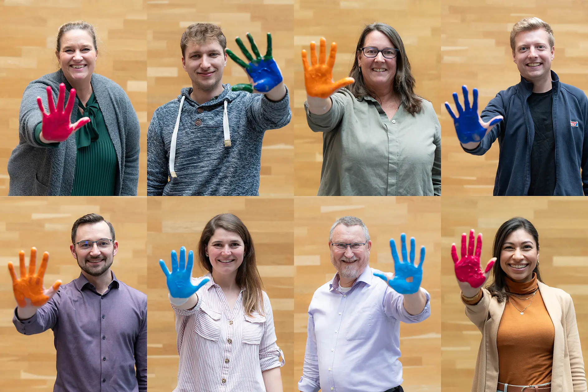 Eight people stand indoors by a light wood wall, holding up hands covered in brightly colored paint, all smiling at the camera.
