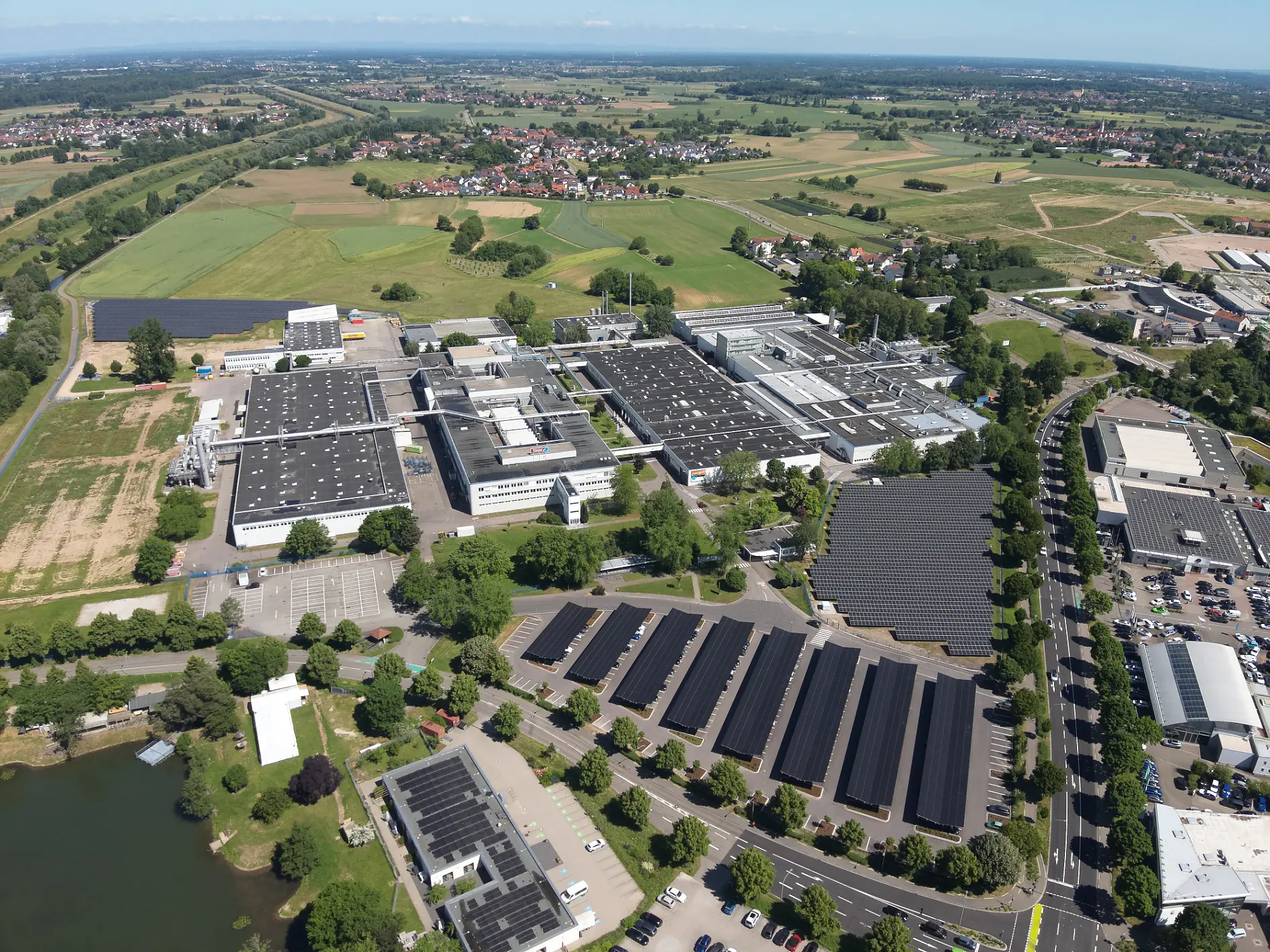 Aerial view of a large industrial complex with tesa tape-used buildings, solar panels on roofs, parking areas, fields, and distant homes. (This text has been generated by AI)