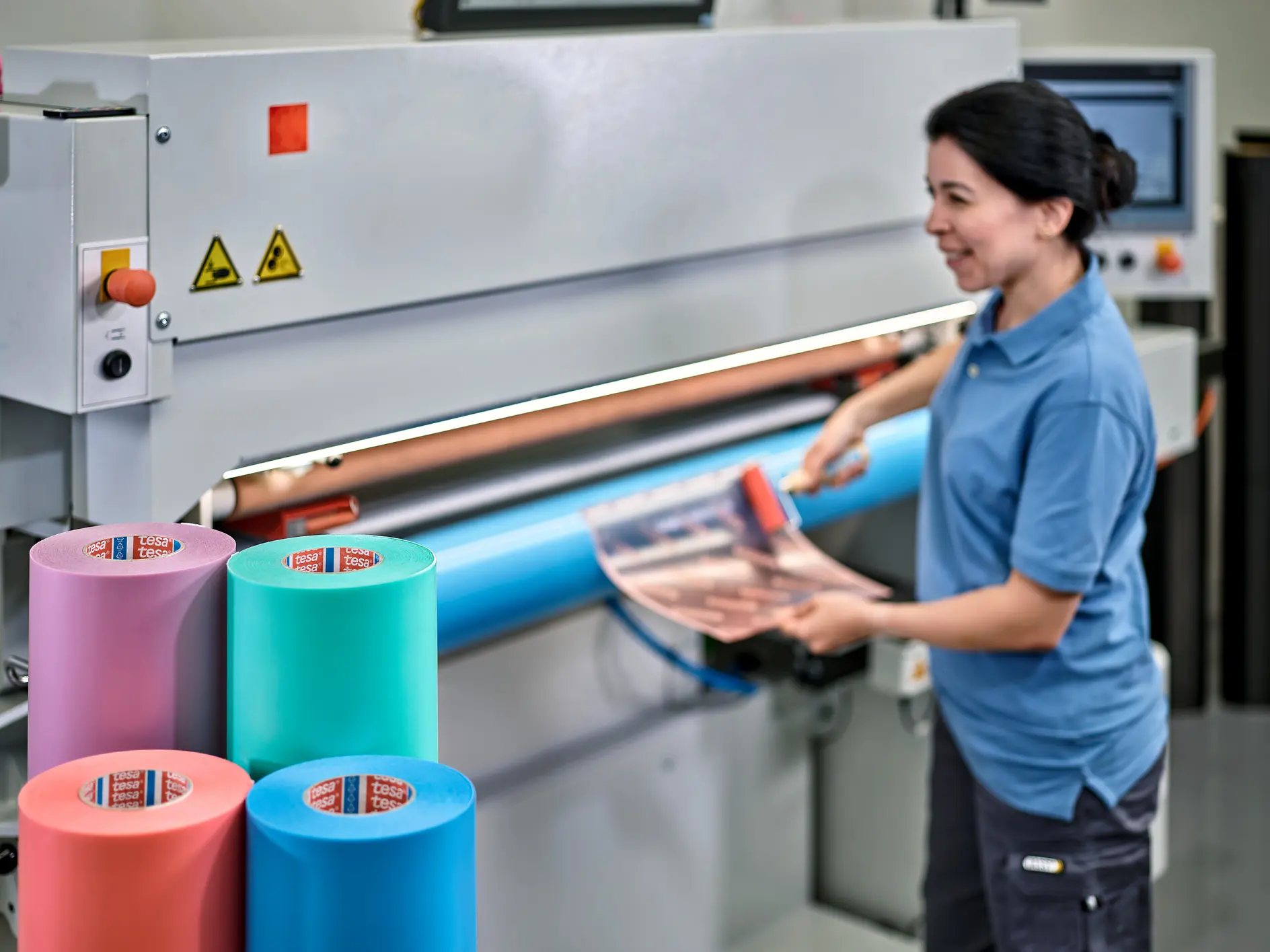 worker operating a large industrial cutting machine indoors with rolls of colorful material labeled tesa nearby (This text has been generated by AI)