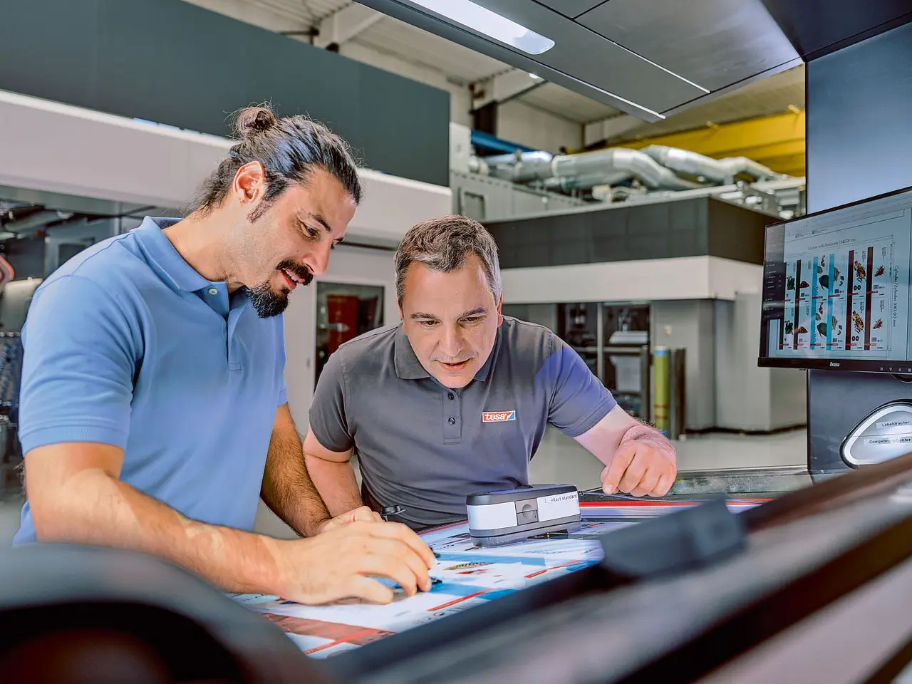 Two men are examining printed pages under a light in an industrial setting. One holds a magnifying glass, while the other points at the paper secured with tesa tape. A computer monitor displaying graphics is visible on the side. Both are wearing casual shirts.