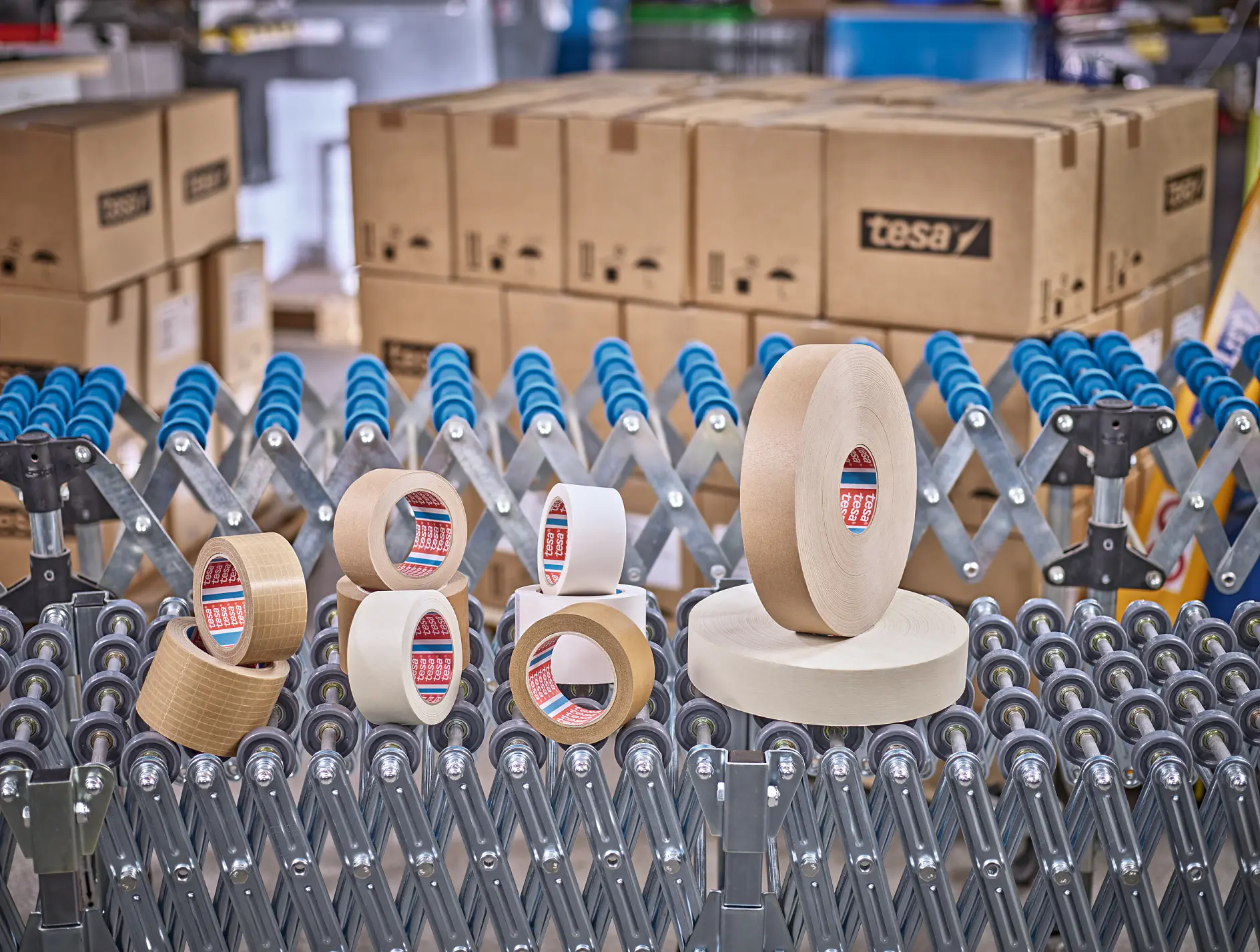 tesa packing tape rolls and dispensers arranged on a metal rack in a warehouse with stacked cardboard boxes in the background (This text has been generated by AI)
