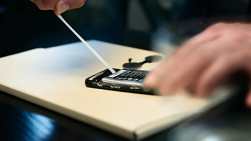 A person is using a plastic tool to remove the back cover of a smartphone placed on a table. The phones battery and internals are visible. The persons hand is slightly blurred, suggesting movement. (This text has been generated by AI)