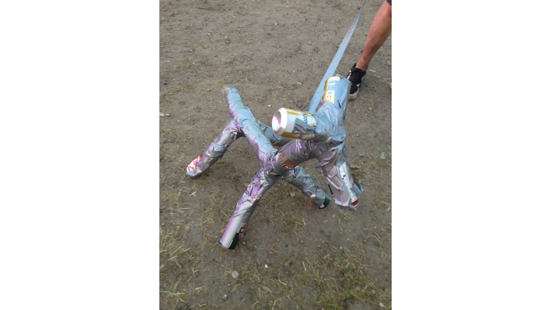 A makeshift sculpture resembling a dog, constructed from aluminum cans and tesa tape, stands on a dirt and grass surface. A persons leg is visible in the background. (This text has been generated by AI)