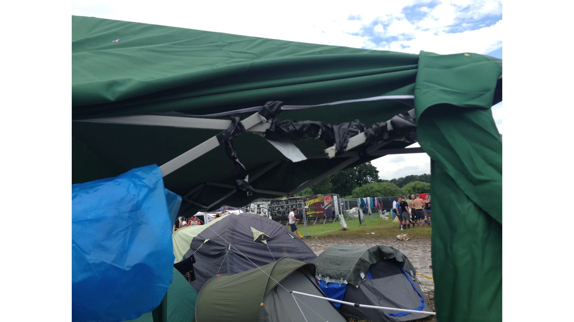 A green tent with a damaged frame secured by black tesa tape, set in a field with additional tents nearby. A group of people stands in the background near a fence under a cloudy sky. The ground appears muddy. (This text has been generated by AI)