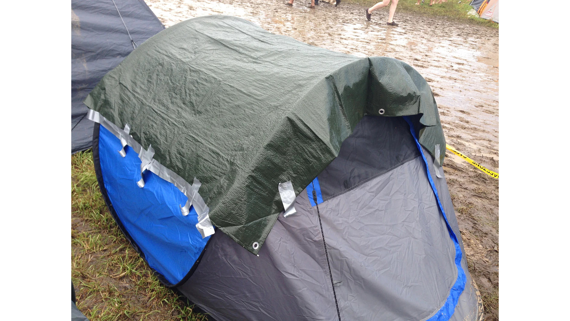 A small tent set up on muddy ground with a green tarp secured over its top. The tarp is held in place with strips of tesa tape. Part of a persons leg is visible in the background. (This text has been generated by AI)