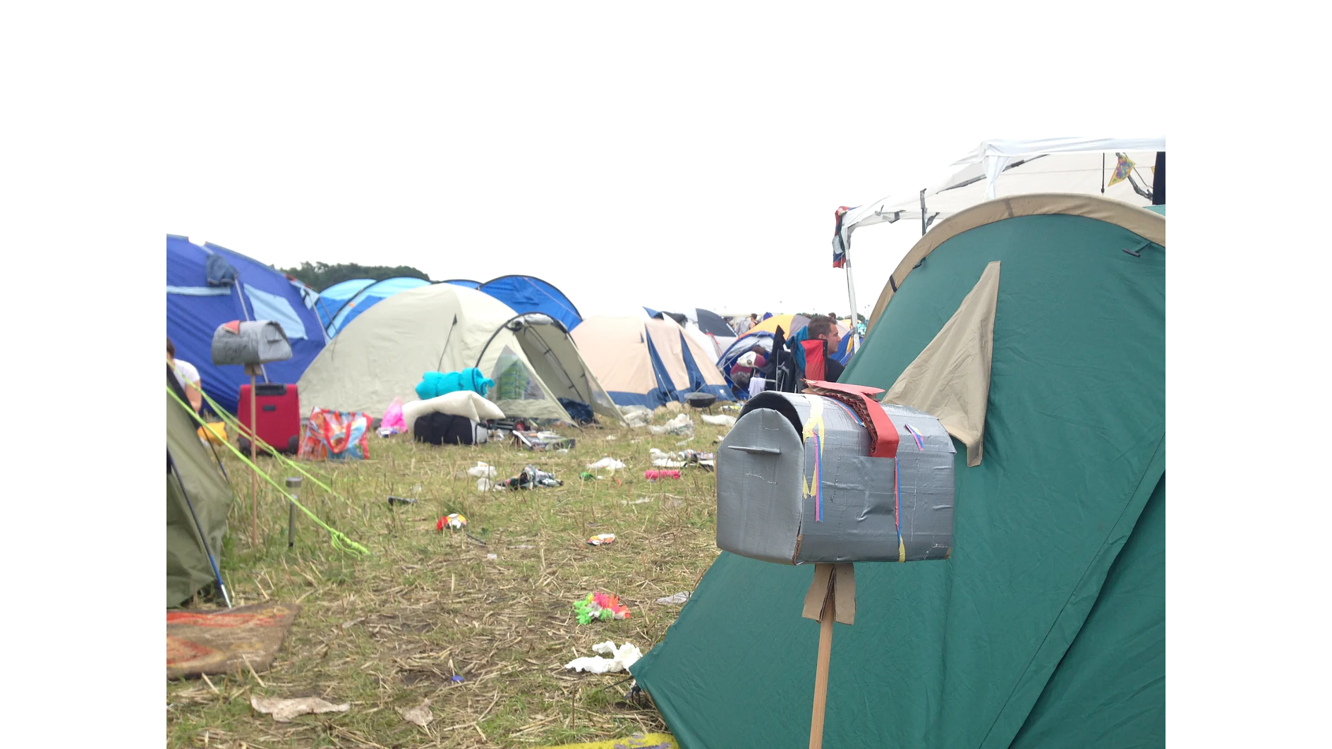 A campsite with several pitched tents of various colors. A makeshift mailbox is attached to a wooden post, covered in tesa tape. The ground is scattered with trash and other camping debris. The sky is overcast. (This text has been generated by AI)