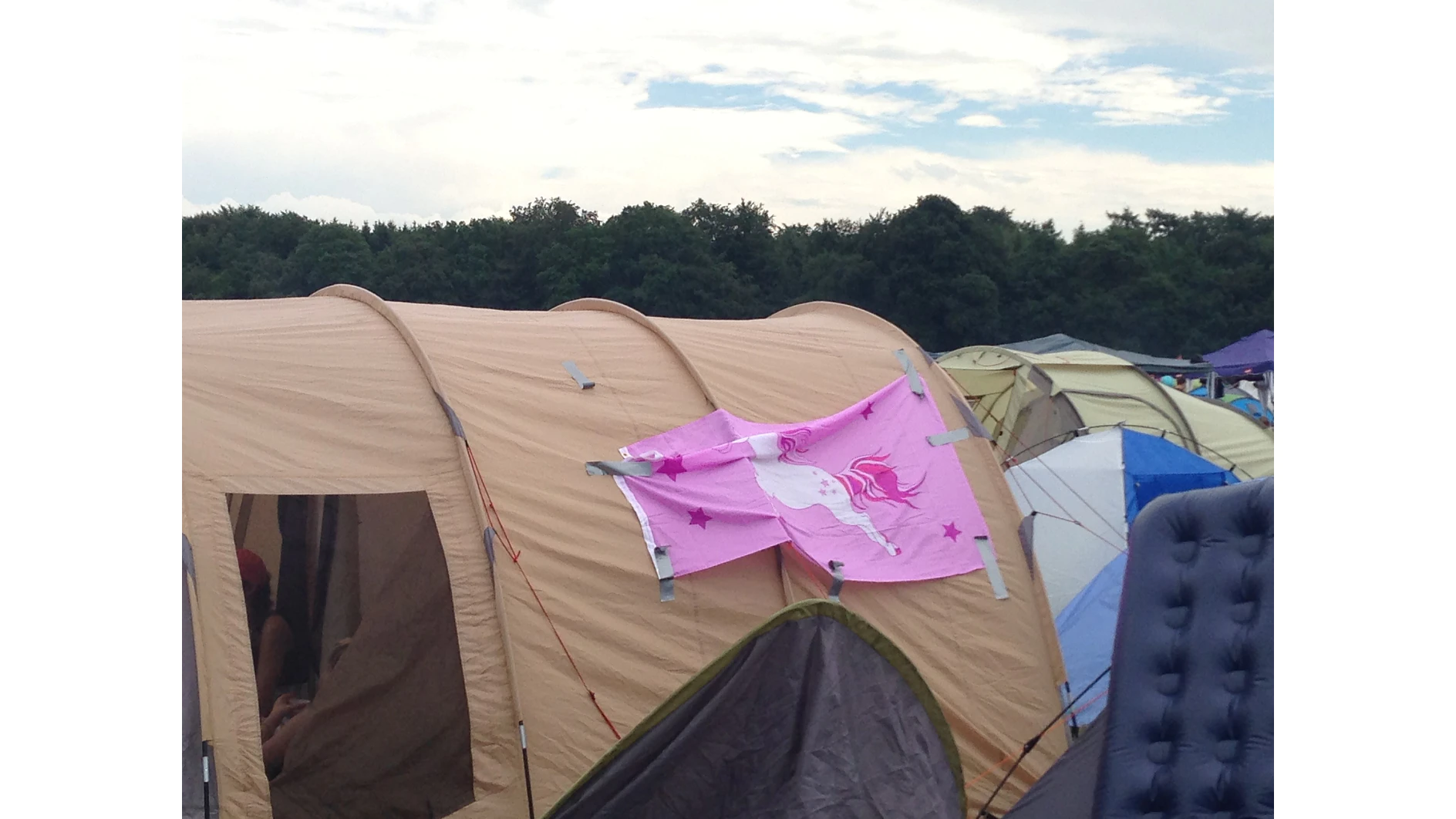 A large beige tent with a pink flag featuring a white unicorn and stars is secured to its side using tesa tape. Several other tents are visible nearby. A forested area is in the background under a cloudy sky. (This text has been generated by AI)