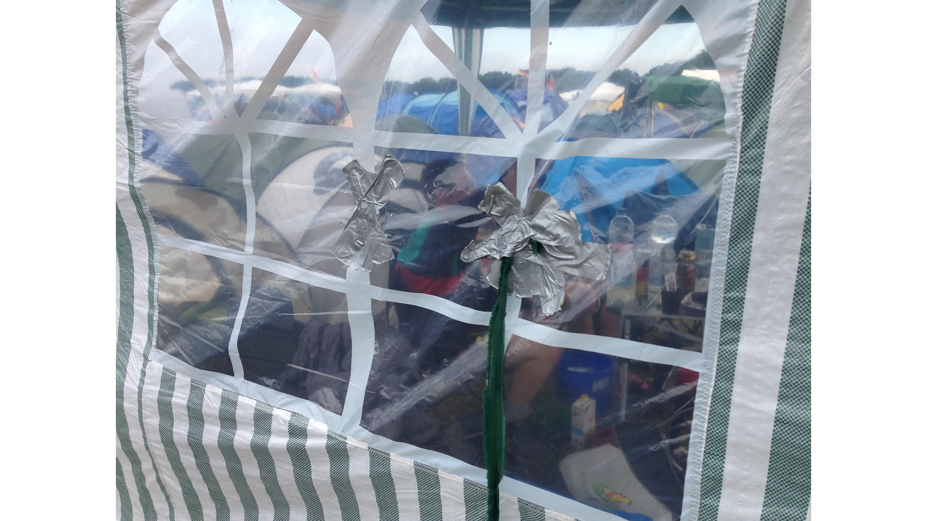 Clear plastic window on a striped tent reveals a view inside, with chairs and multiple colorful tents visible in the background. Two pieces of tesa tape form an X and a flower shape on the window. (This text has been generated by AI)