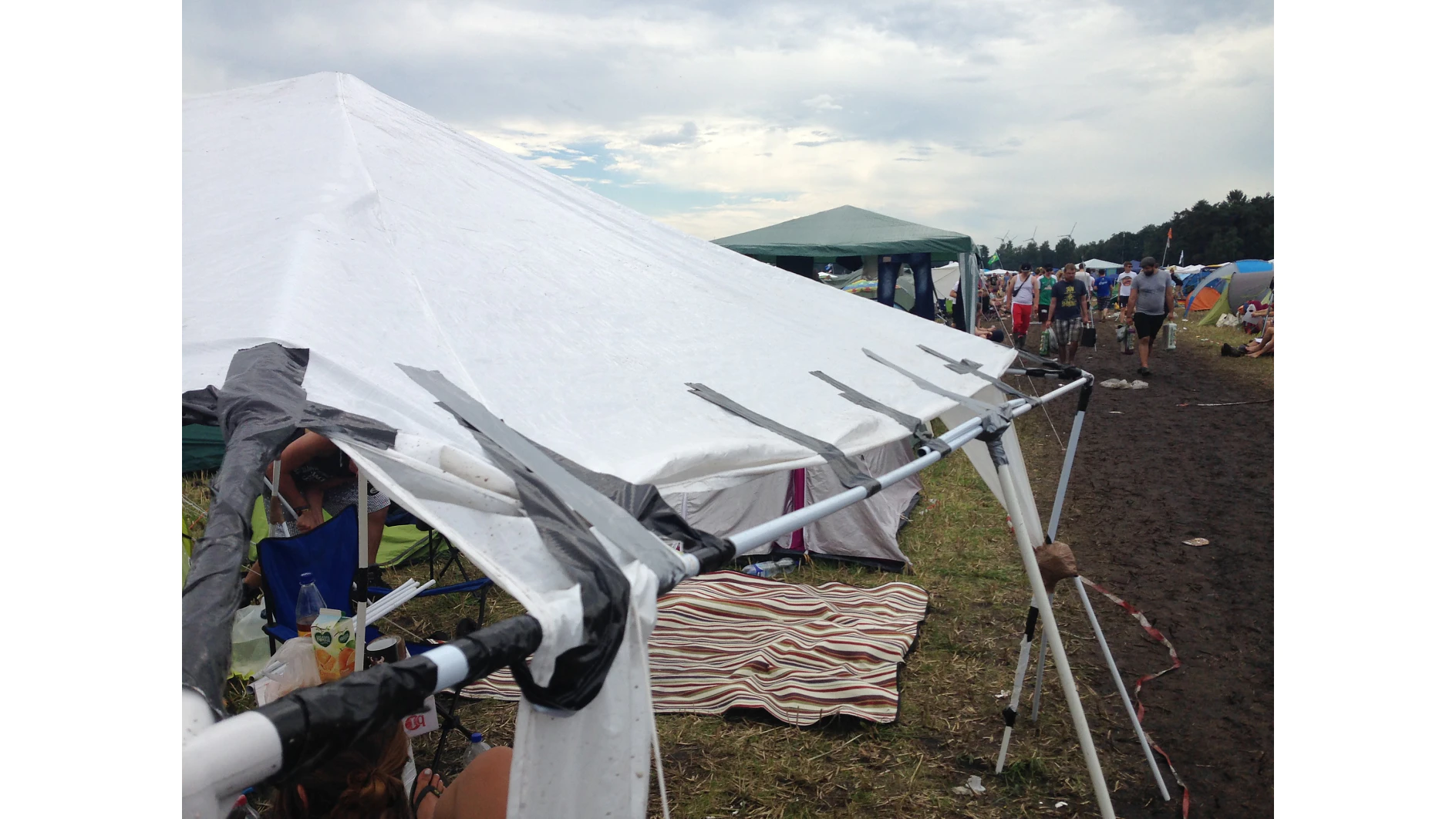 A makeshift tent with a white tarp secured by black tesa tape to a metal frame at an outdoor event. People are in the background, and the ground is muddy with scattered grass. A striped blanket and some camping items are partially visible. (This text has been generated by AI)