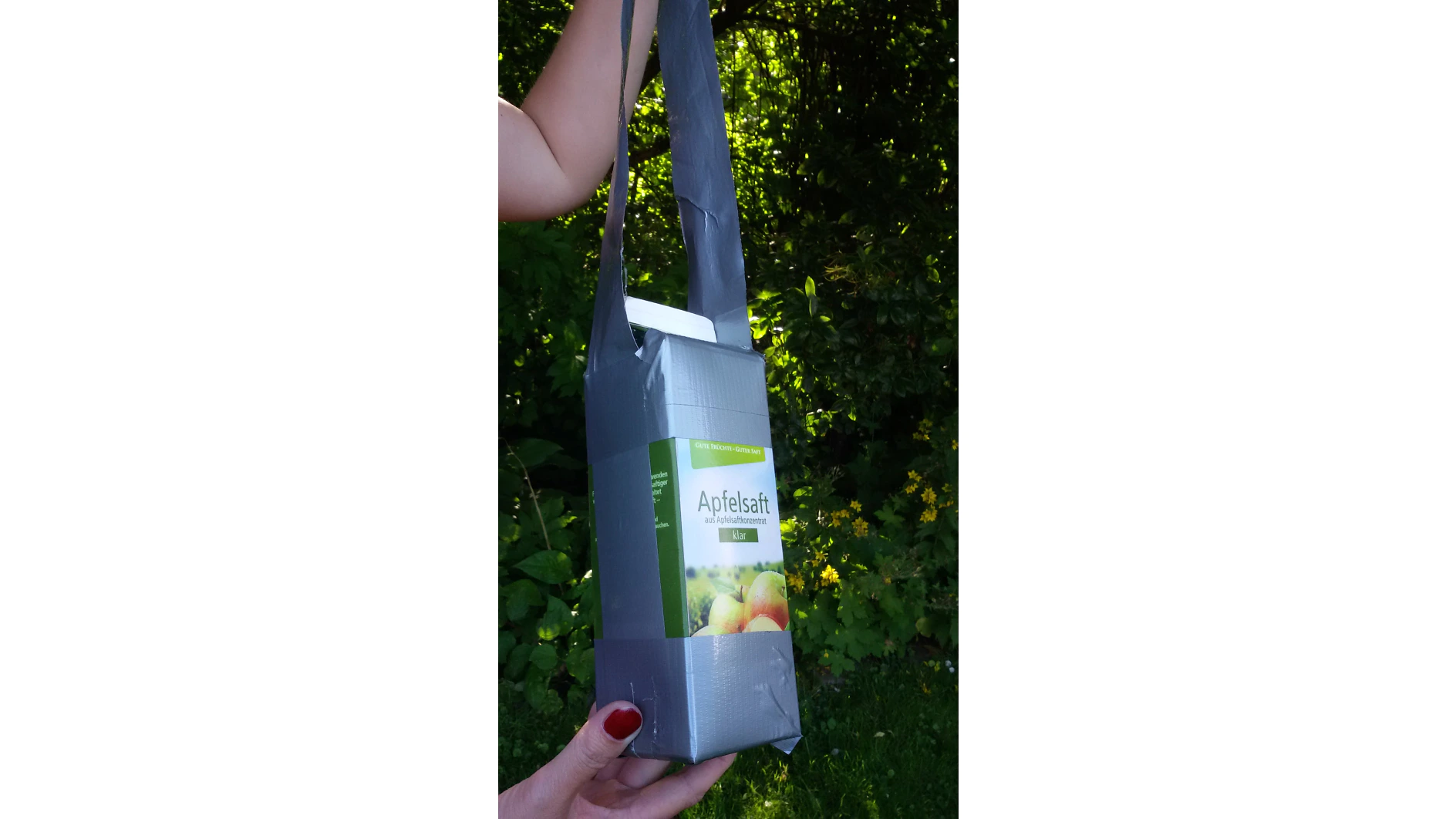 A hand with red nail polish holds a carton of apple juice with a handle made from grey tesa tape. The carton is labeled Apfelsaft. The background is filled with green foliage and yellow flowers. (This text has been generated by AI)