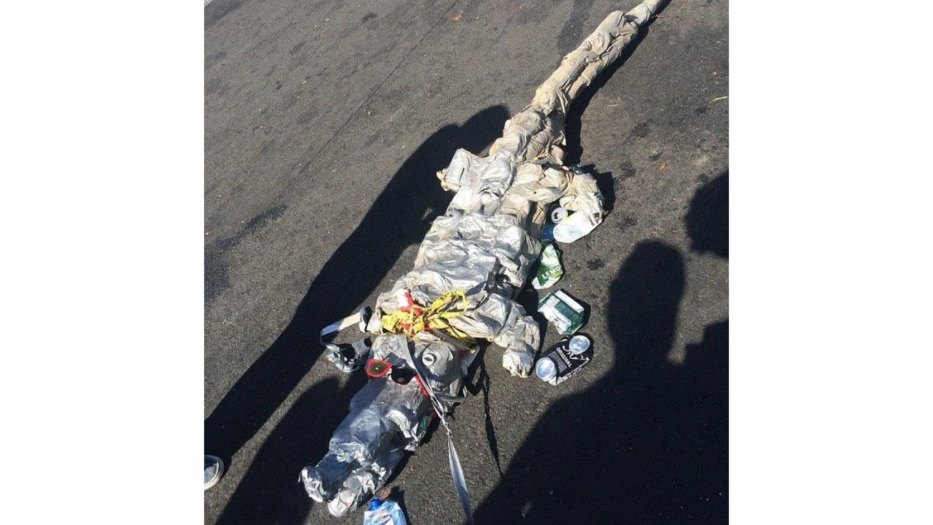 A sculpture of a crocodile made from crushed cans and cardboard lies on a pavement. Shadows of people can be seen cast on the ground around it. (This text has been generated by AI)