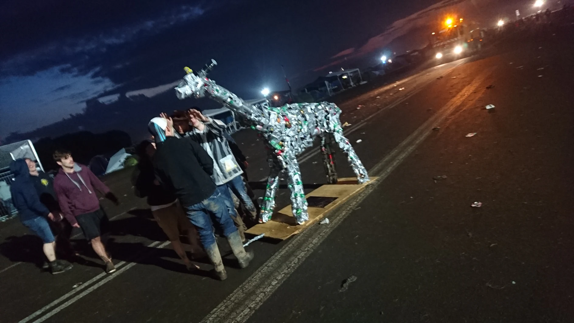 A group of people stand near a giraffe-shaped sculpture made from recycled materials on a road. The scene is set at dusk with scattered debris and tents in the background. Bright lights illuminate the area, highlighting the creative use of tesa tape throughout the artwork. (This text has been generated by AI)