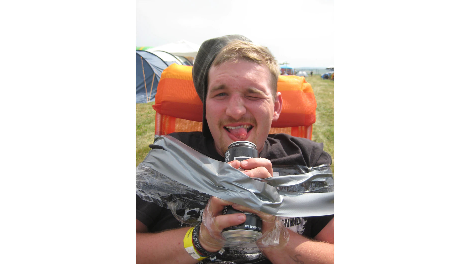 A person sits in an orange chair outdoors, playfully sticking their tongue out while holding a beverage can wrapped in plastic and tesa tape. They are wearing a dark shirt and a cap, with tents visible in the background. (This text has been generated by AI)