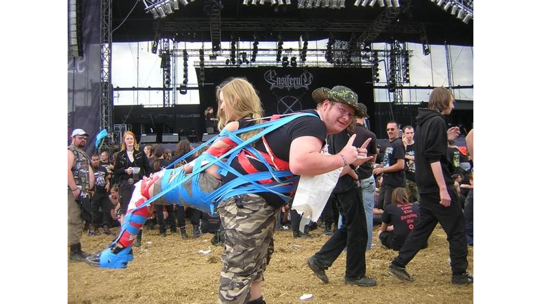 A person is attached to anothers back using blue and red tesa tape at an outdoor event. The taped person is wearing camo pants. The background showcases a stage and a crowd standing on straw-covered ground. The person in front is making a hand gesture. (This text has been generated by AI)