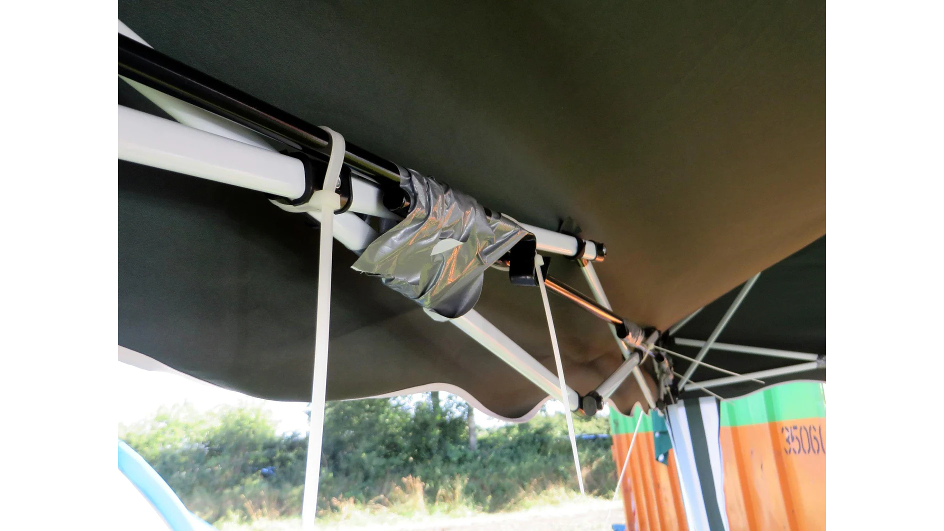 A canopy frame with a section reinforced by tesa tape is shown. The tesa tape secures two metal poles together. The canopy fabric is dark green and covers the frame. The background includes blurred greenery and part of a colorful striped object. (This text has been generated by AI)