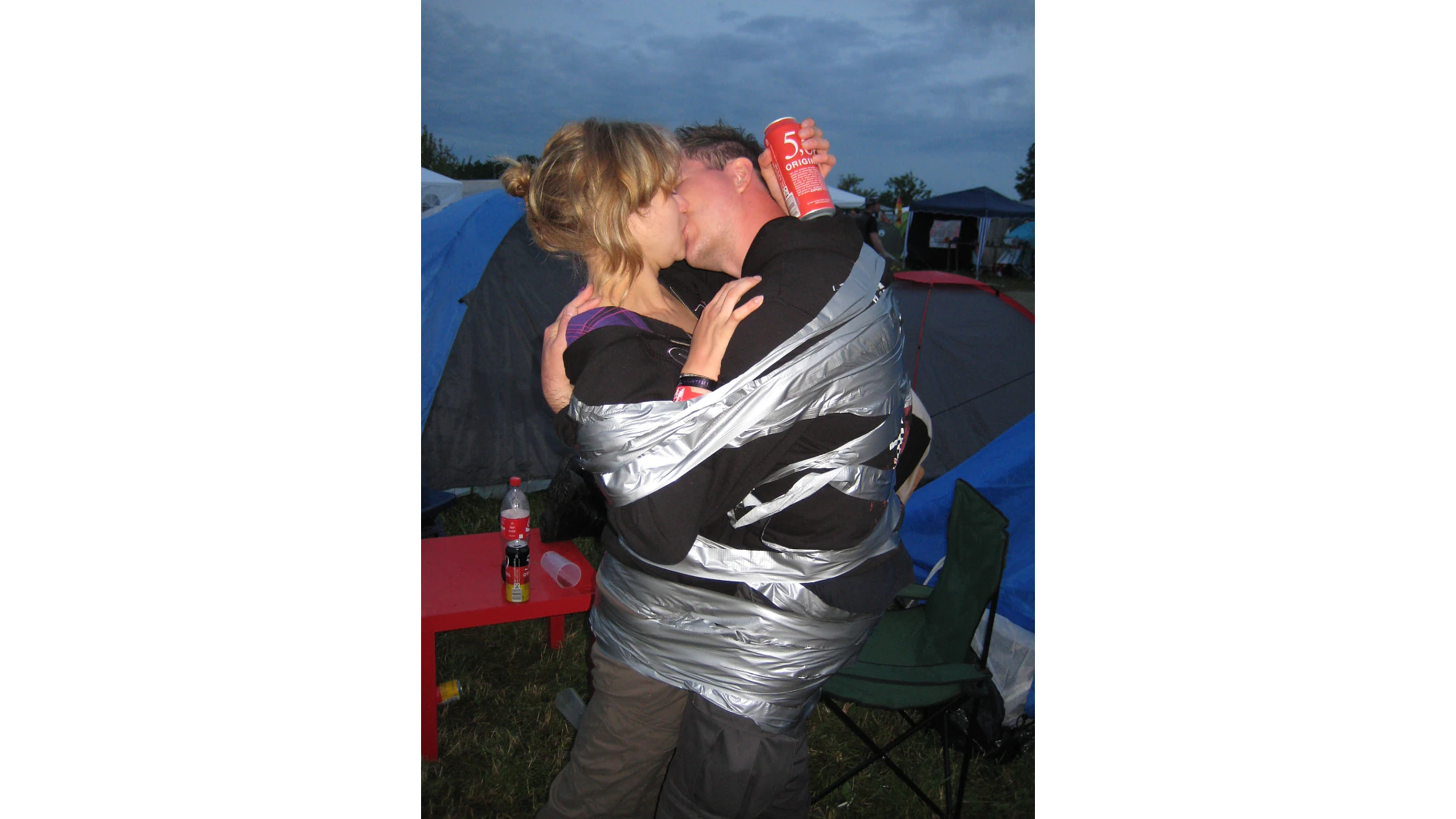 Two people are wrapped together with silver tesa tape, sitting in a camping chair and kissing. The person on the right holds a red can. Tents and a cloudy sky are in the background, and a small red table with drinks is nearby. (This text has been generated by AI)